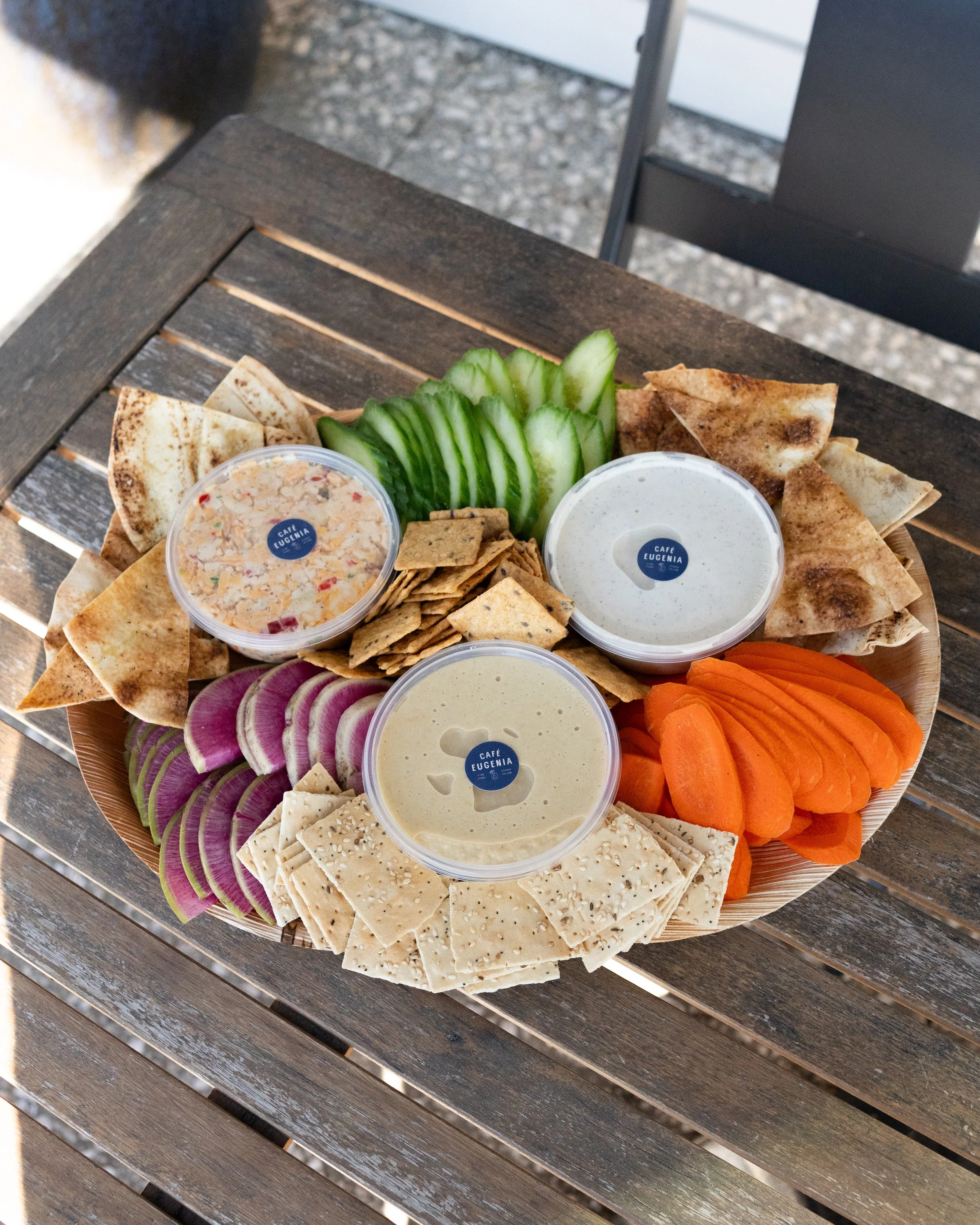A large round wooden platter with sliced vegetables, crackers, flatbread, and three small bowls of sauces, on a wooden table.