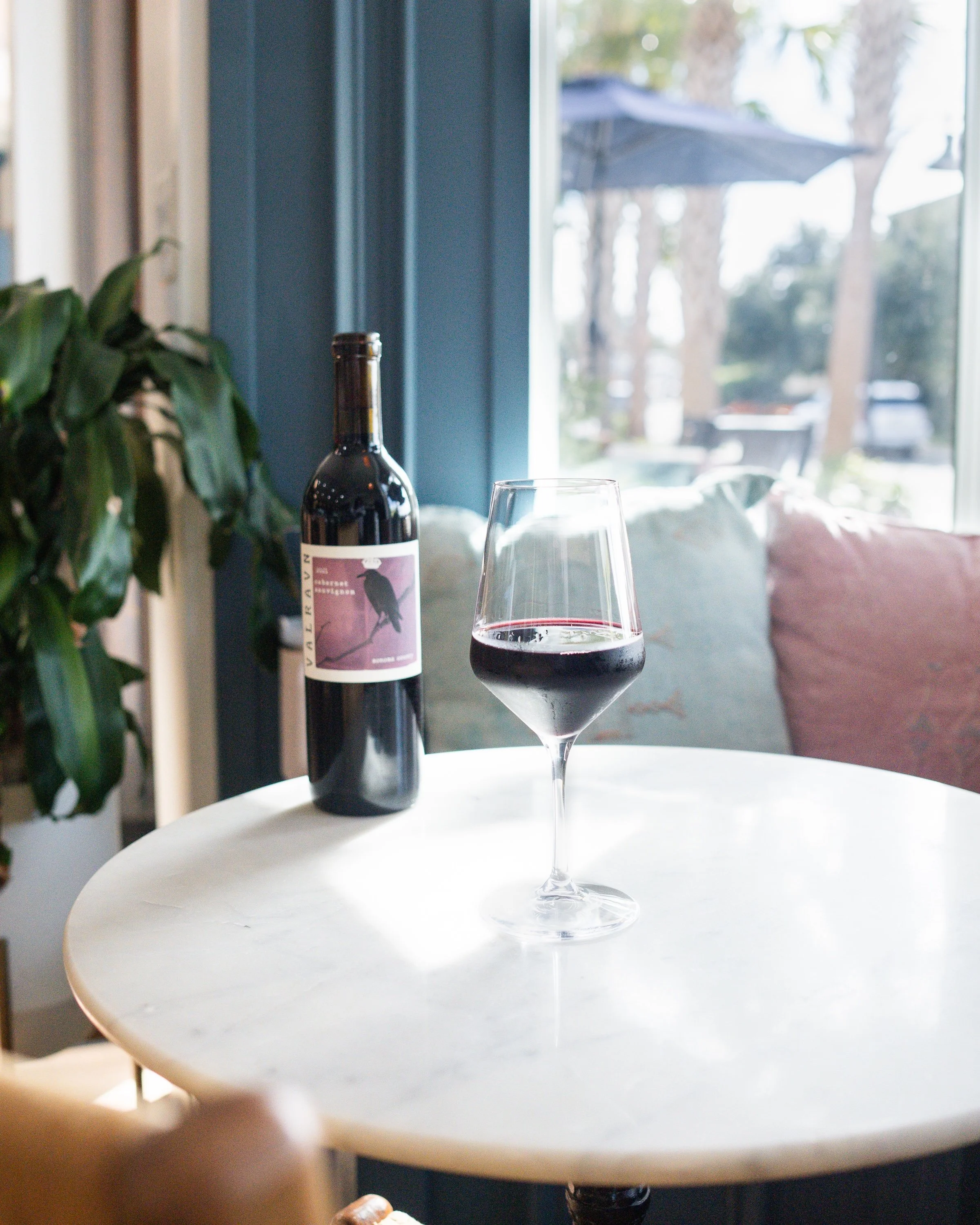 A bottle of red wine and a glass of red wine on a white round table in a cozy room with a large window, outdoor greenery, and cushions on a sofa in the background.