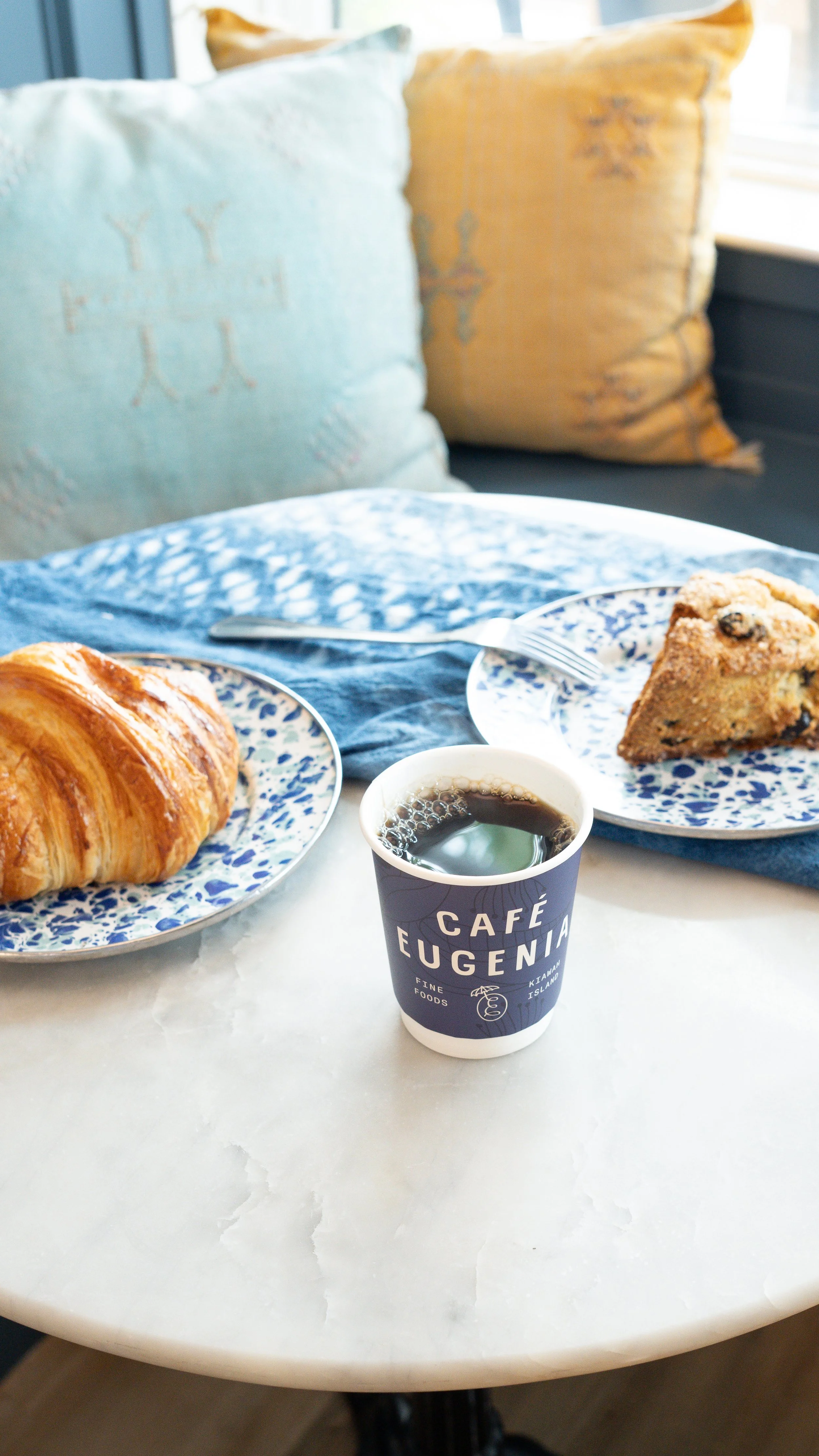 A table with a croissant, a slice of blueberry muffin, and a cup of coffee at Café Eugenia in Kiana, Alaska, with pillows visible in the background.