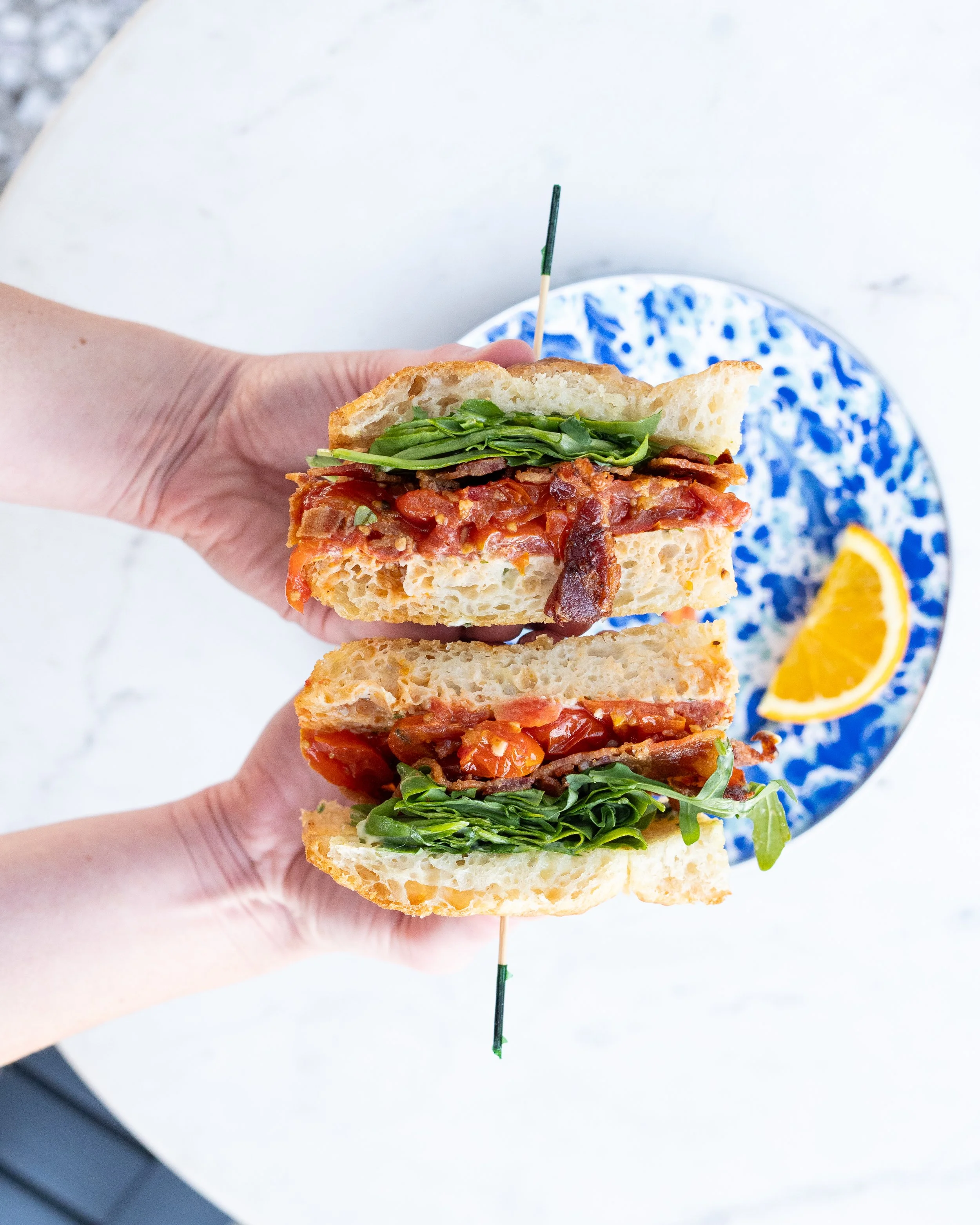Person holding a sliced eggplant parmesan sandwich with basil, tomato sauce, and cheese on a blue and white plate with lemon wedge.