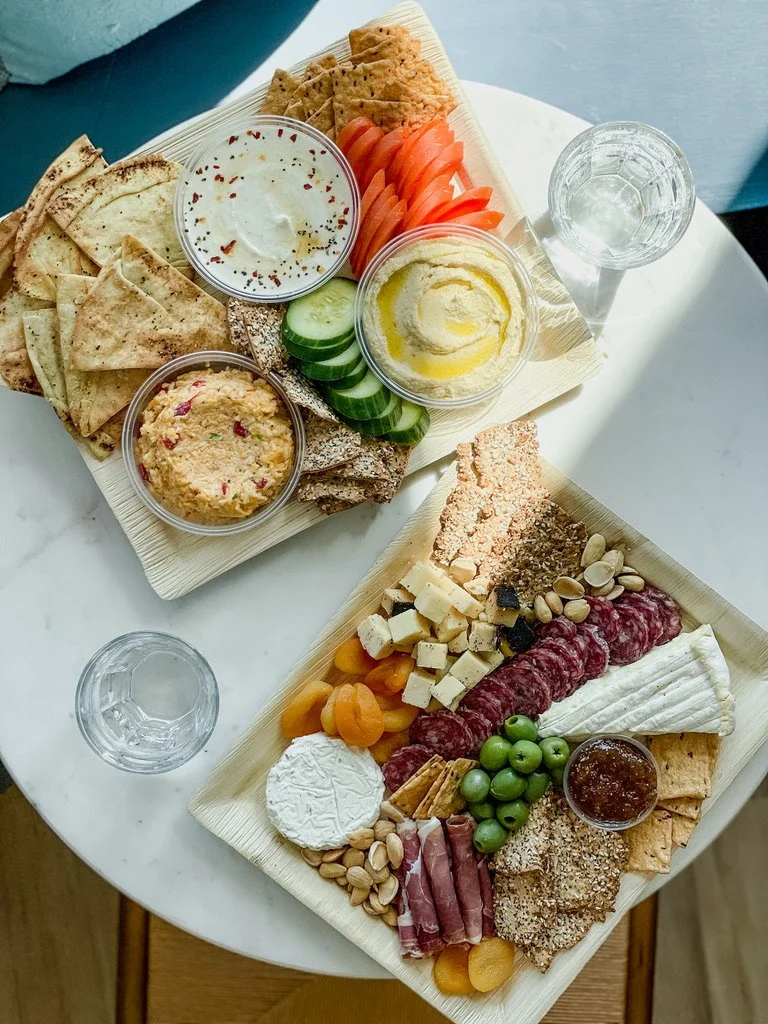 A platter of sliced vegetables, hummus, pita chips, and dips, alongside a cheese, meat, and fruit tray with nuts and a small jar of jam.