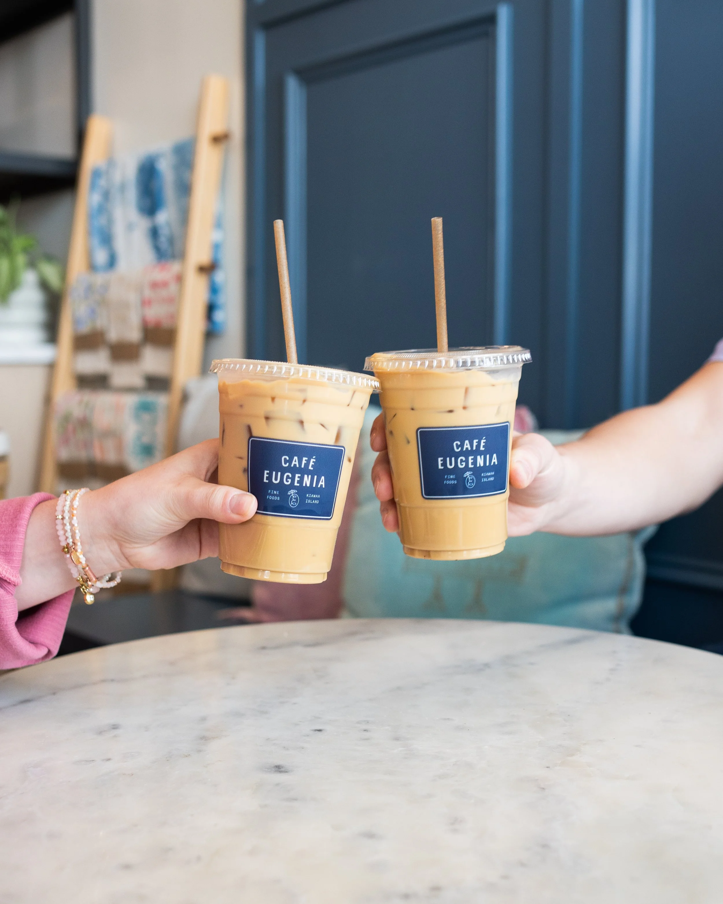 Two hands holding iced coffee drinks with blue labels that read 'Café Eugenia.'