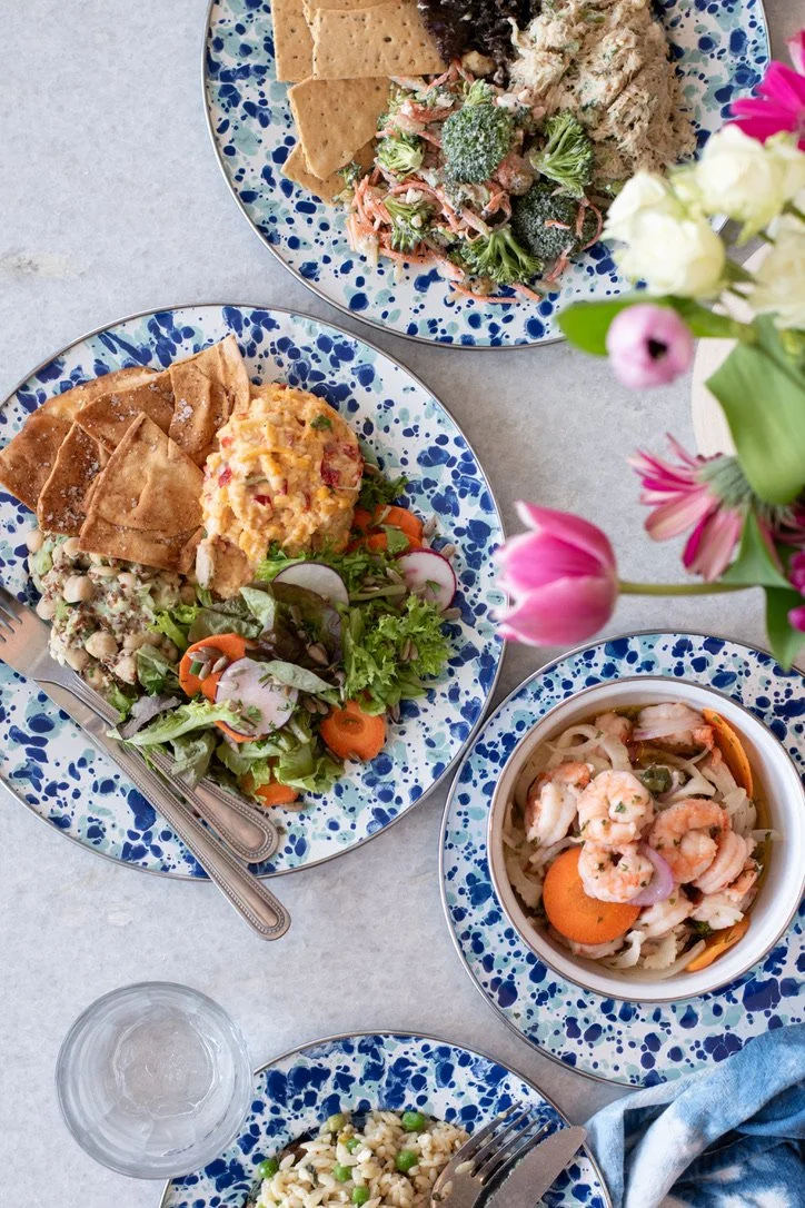Various colorful plates of salads and dishes with a vase of pink flowers on a light-colored table.