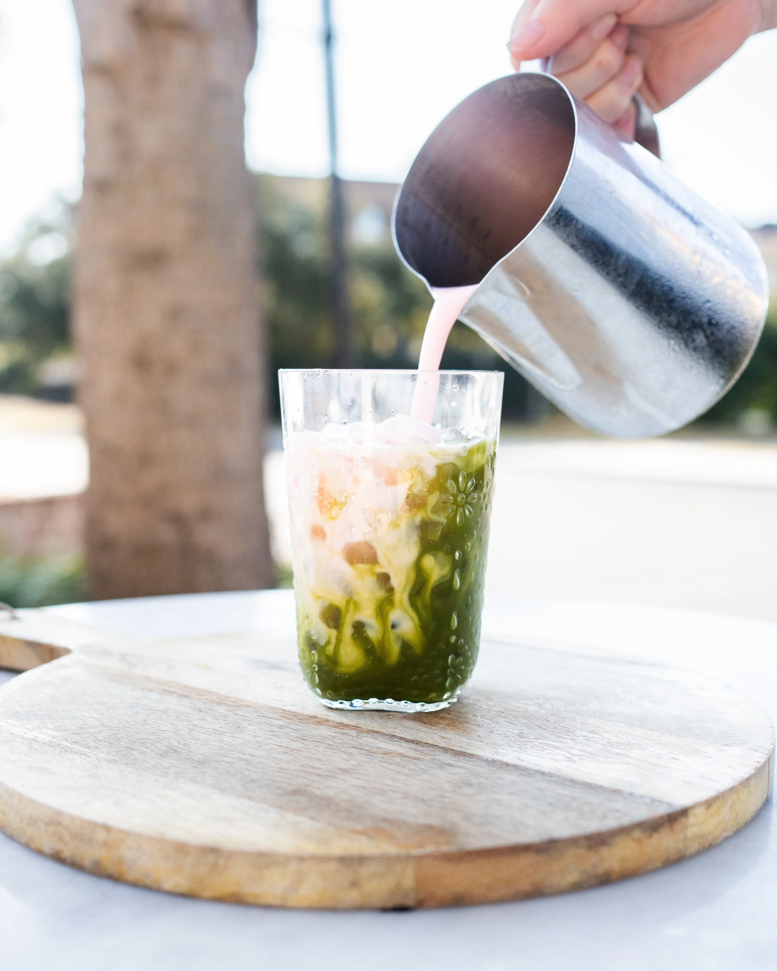 A glass with layered matcha and milk, with pink milk being poured into it from a metal pitcher, on a wooden board outdoors.