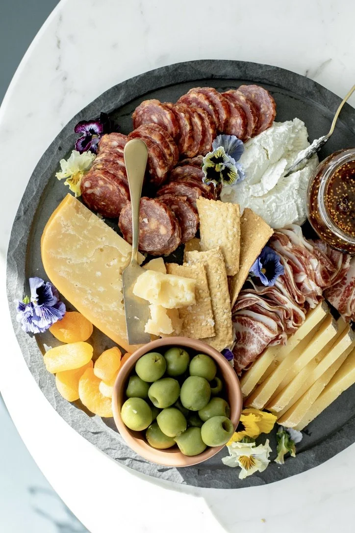 Charcuterie board with various cheeses, sliced cured meats, green olives, dried apricots, and edible flowers on a marble surface.