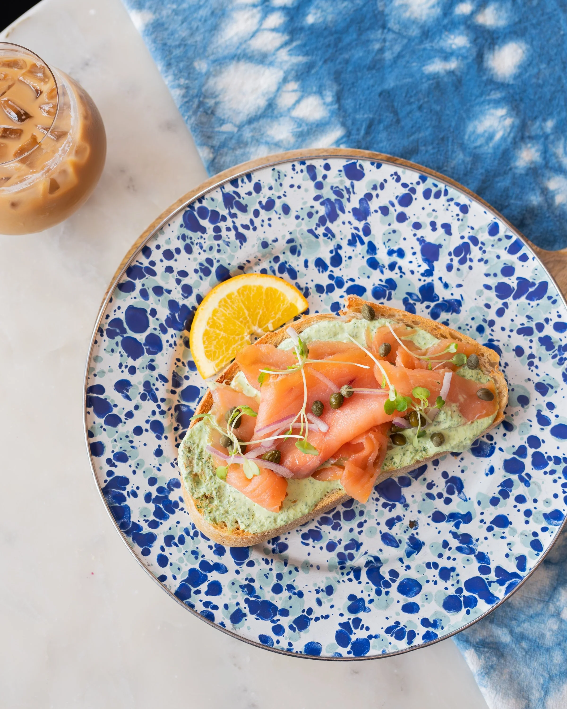 Smoked salmon and avocado toast garnished with microgreens and capers, served with a lemon slice on a blue speckled plate.