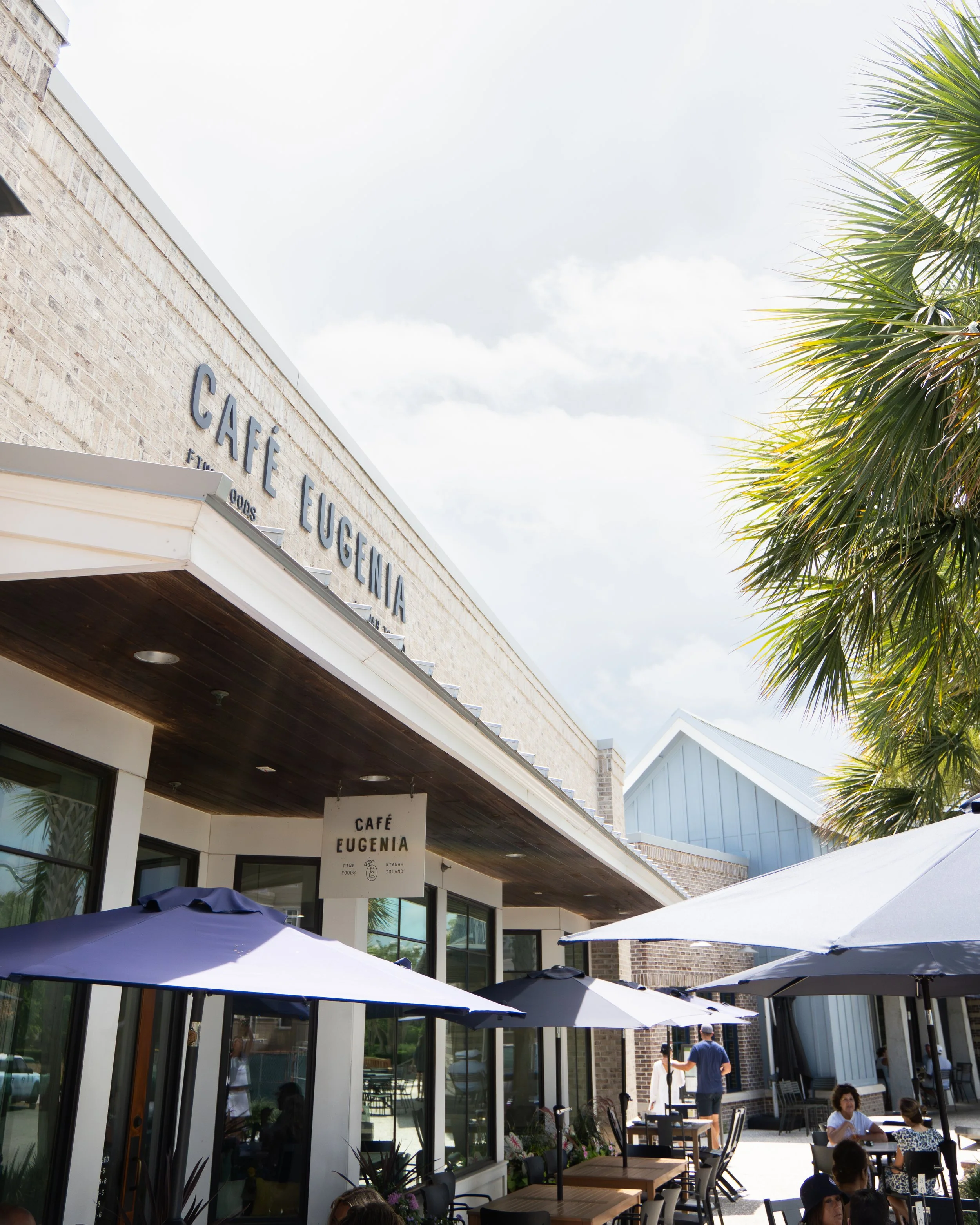 Outdoor seating area of Café Eugenia with tables, chairs, and umbrellas, adjacent to a building with a sign reading 'Café Eugenia' and palm trees.