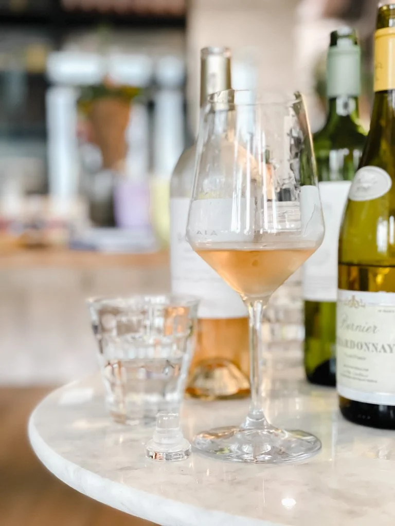 A glass of rosé wine, a small glass of water, and several bottles of wine on a marble table in a cozy setting.