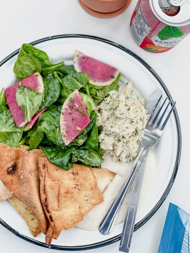 Salad with spinach and watermelon radishes, a serving of tuna salad, pita bread, and a side of yogurt or cream sauce on a white plate, with a fork, can of juice, and additional food items nearby.