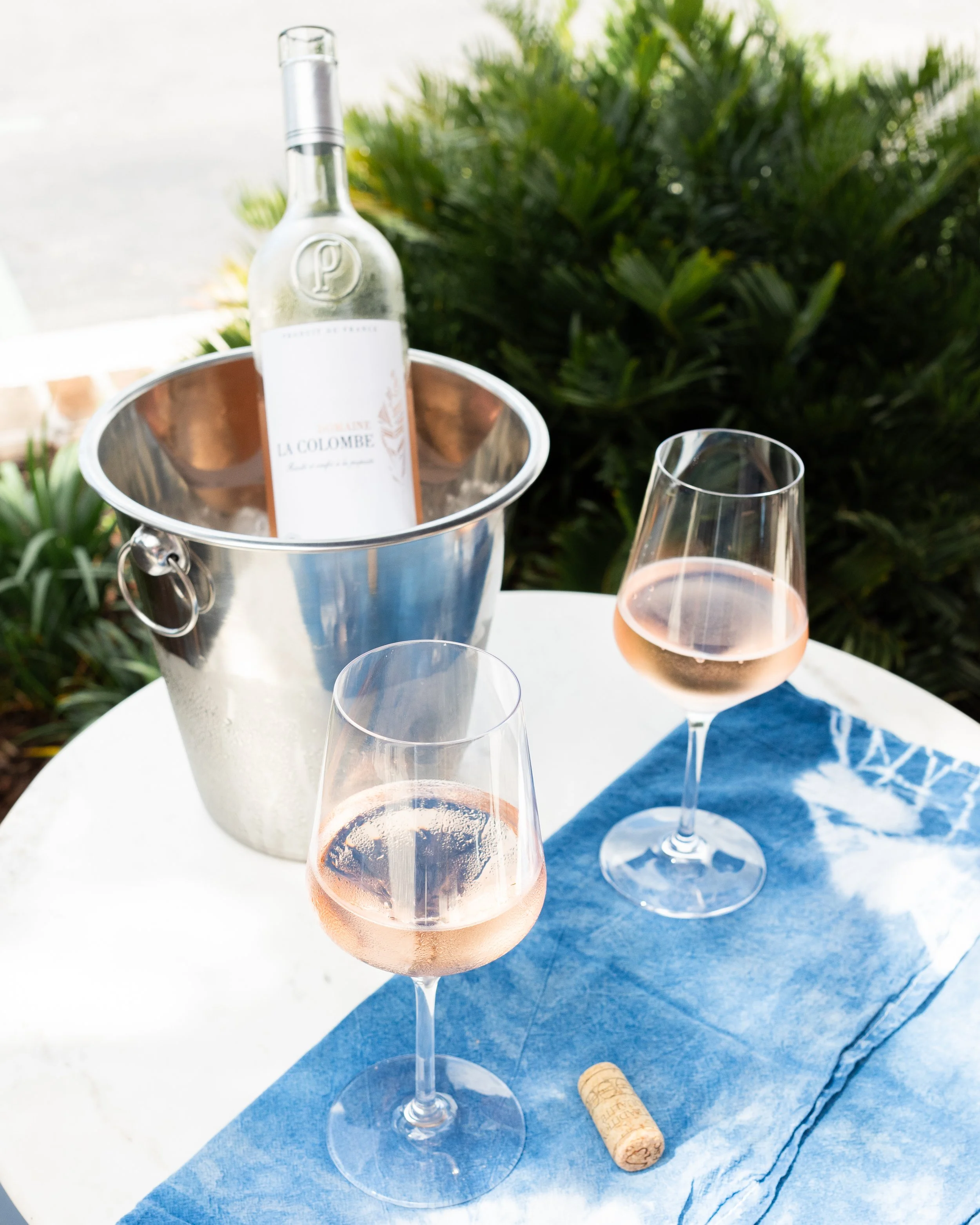 Two glasses of rosé wine on a white table with a blue cloth, a cork, and a bottle of rosé in a metal ice bucket, surrounded by greenery.