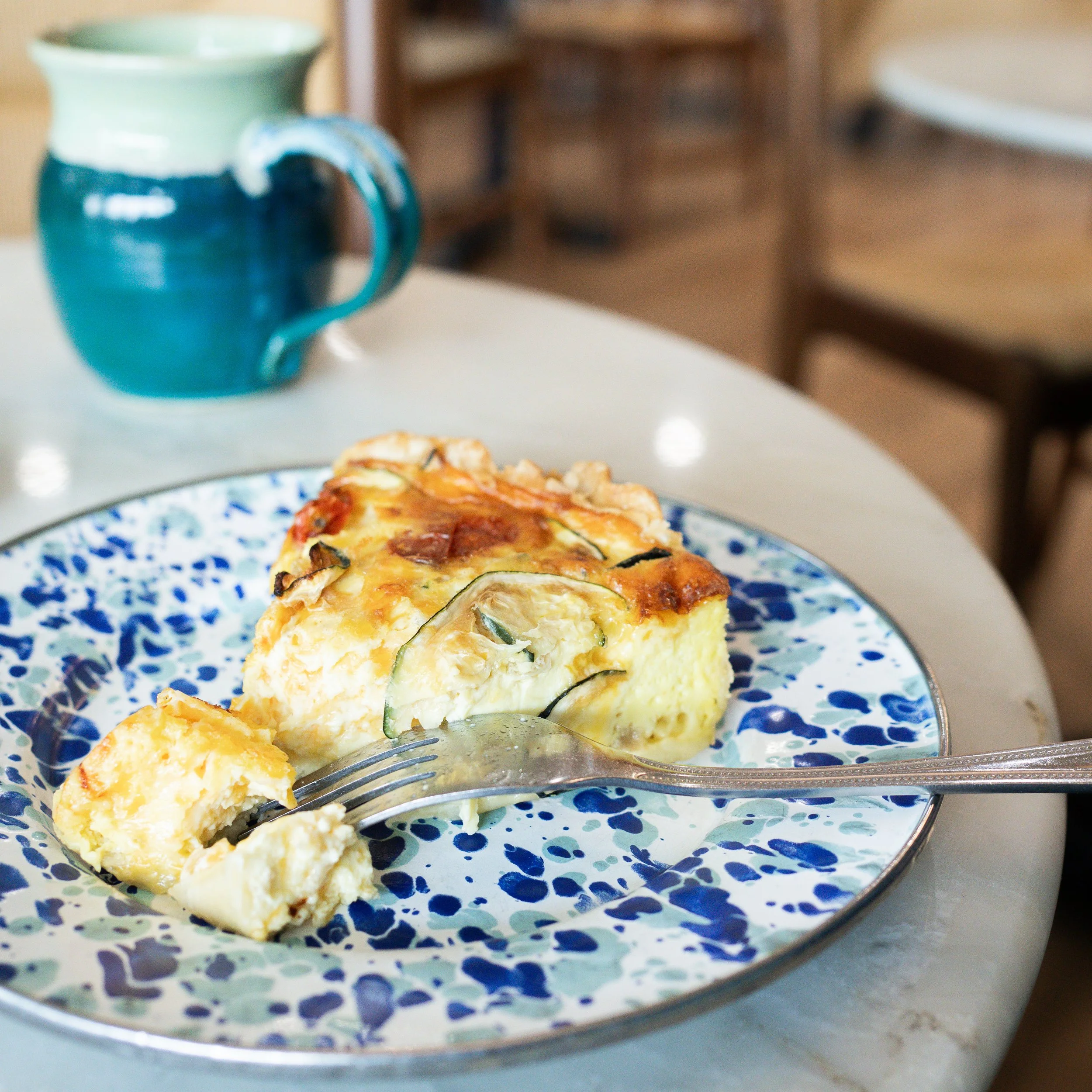 A slice of zucchini quiche on a blue and white patterned plate with a fork, placed on a white table, with a blue and green ceramic mug in the background.