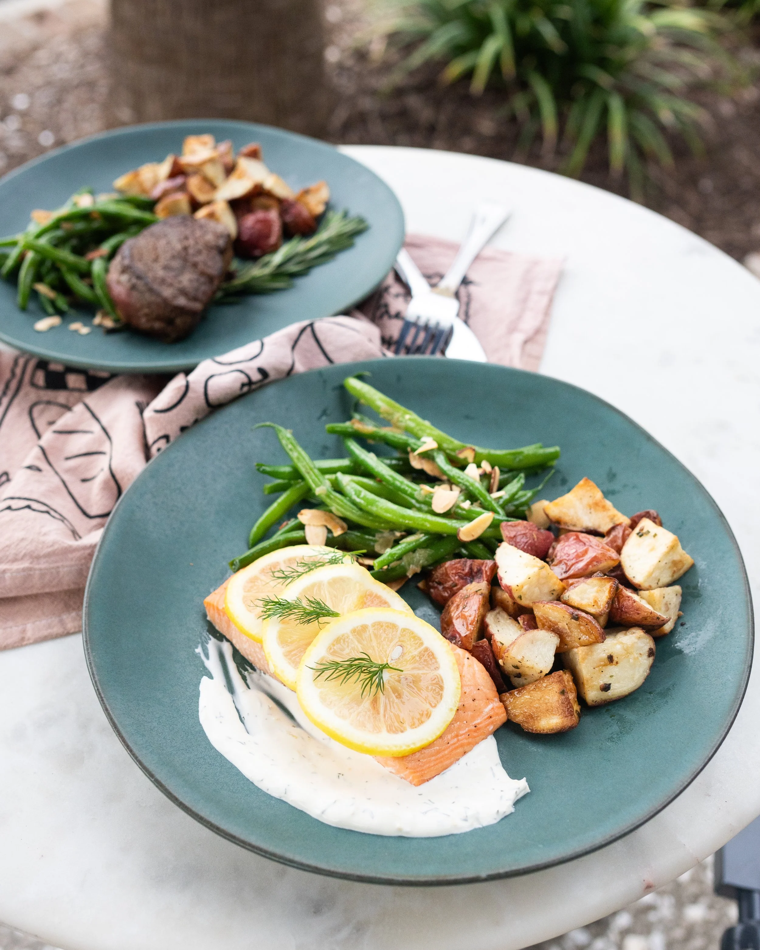 Plate with salmon topped with lemon slices and dill, green beans, roasted potatoes, and sauce on a white marble table.