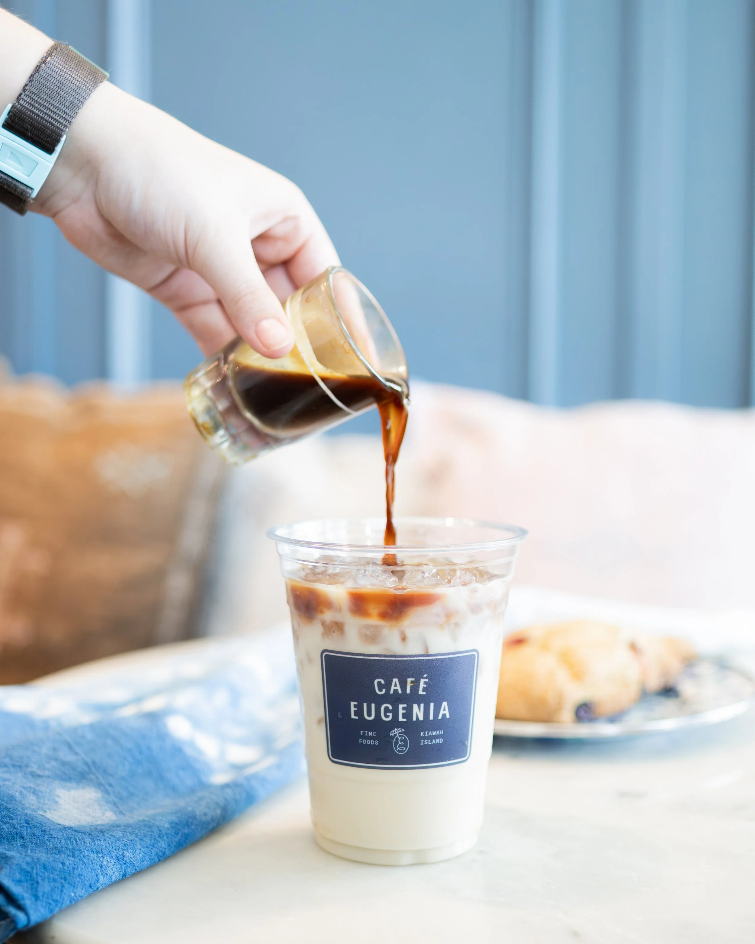 A hand pouring coffee from a small glass pitcher into a plastic cup of iced coffee on a table, with a plate of pastry in the background.