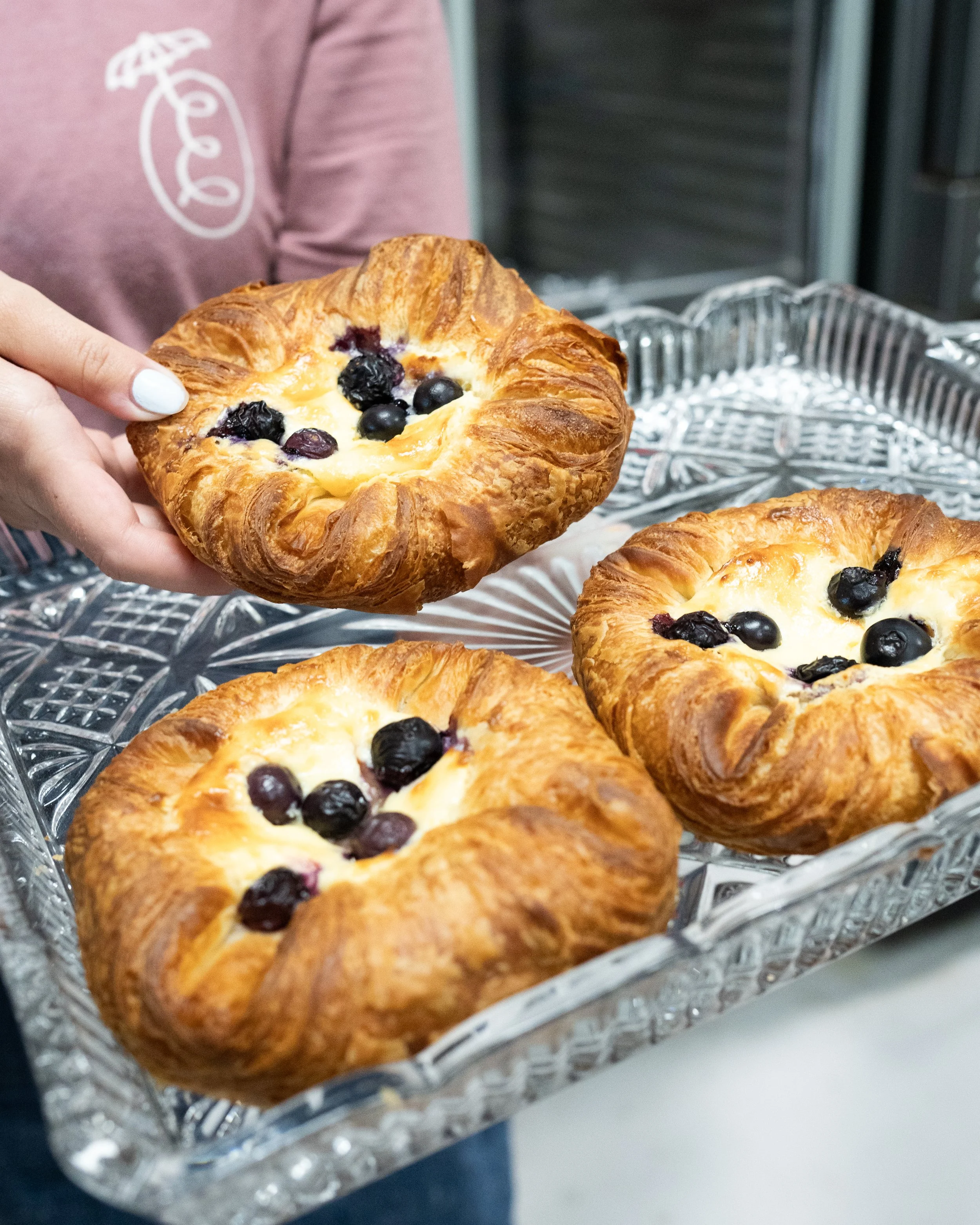 Person holding a blueberry Danish pastry over a glass tray with three more blueberry Danish pastries.