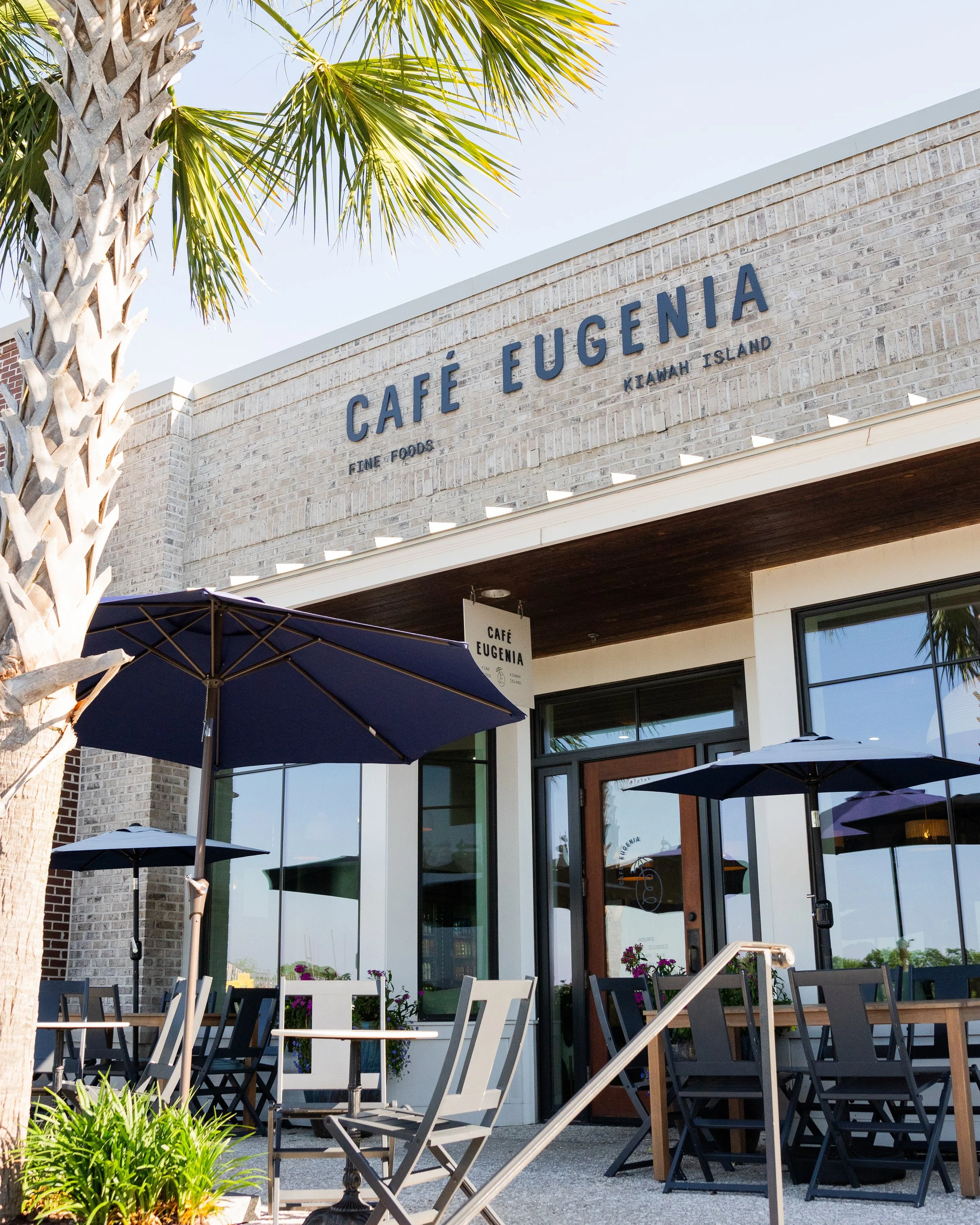 Exterior view of Café Eugenia with outdoor seating, umbrellas, and palm trees.