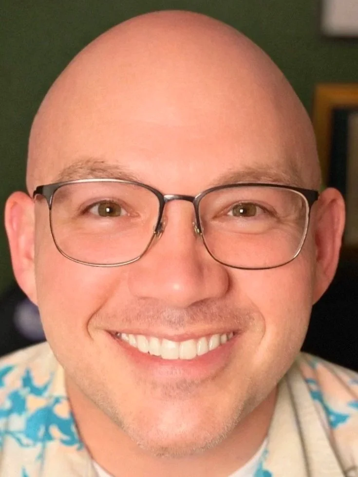 A smiling man with glasses, light skin, no hair, and wearing a colorful shirt.