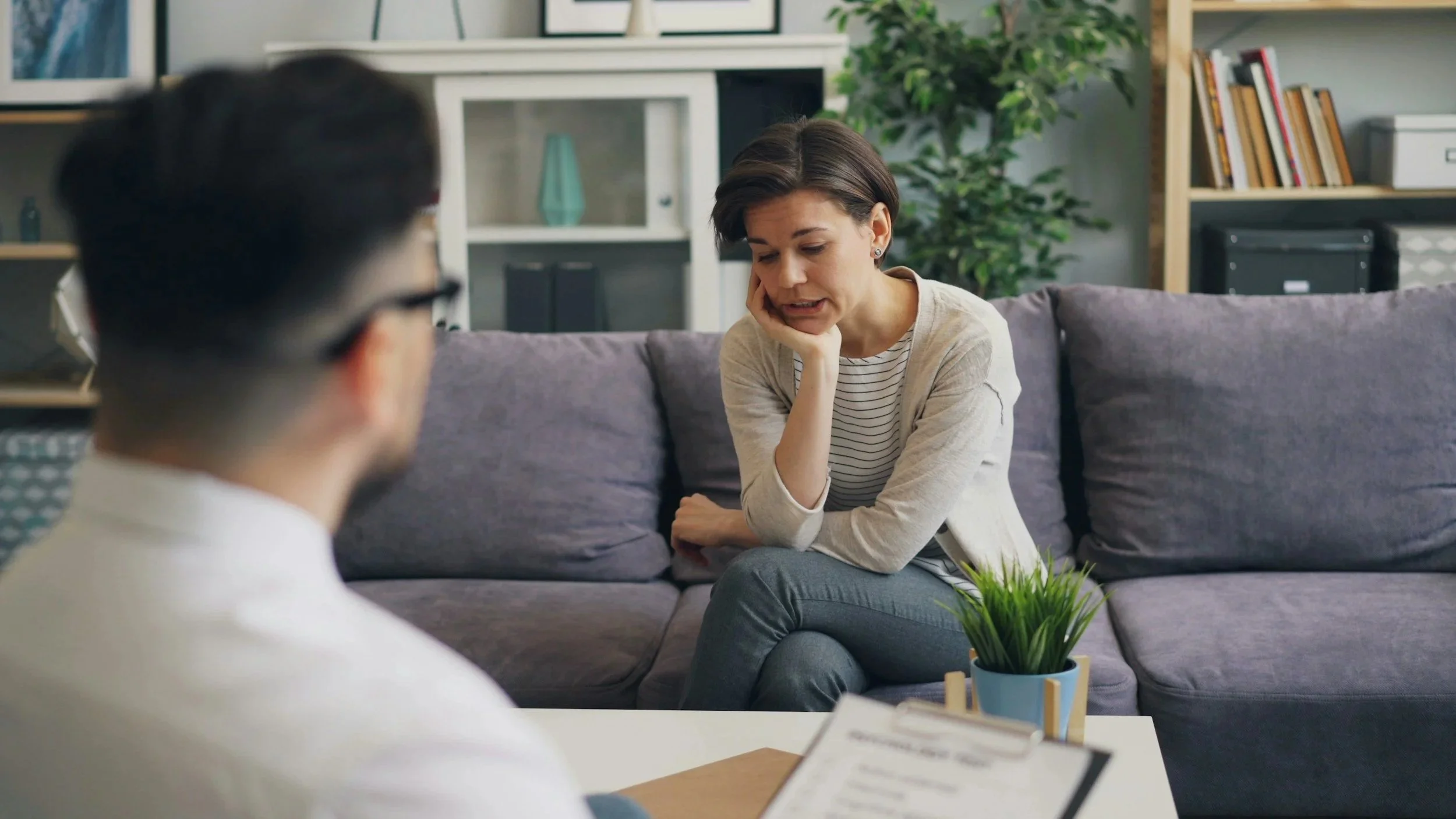 A woman sitting on a gray couch with a worried expression, talking to a therapist or counselor. The therapist, visible from the back, is wearing glasses and a white shirt. There is a small potted plant on a white table in front of them, and background shelves with books and decor items in a cozy, well-lit room.