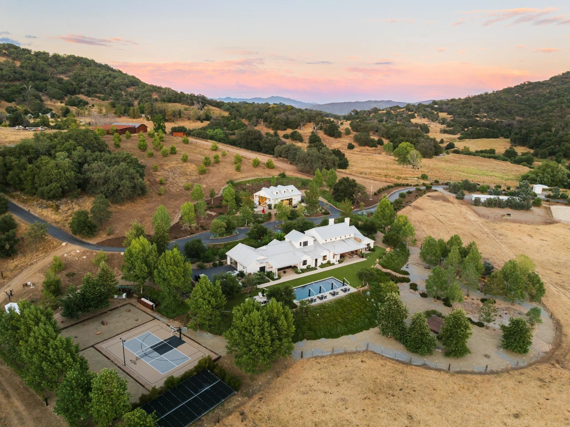 Aerial view of a large countryside estate with a white house, swimming pool, tennis court, and surrounding trees, hills, and farmland at sunset.