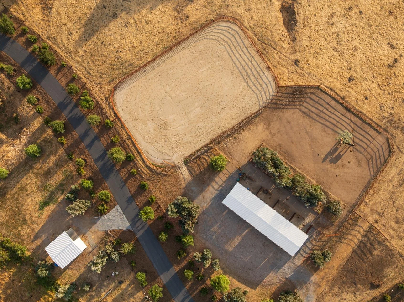 An aerial view of a sports complex with two fenced sports courts, a parking area, a building, trees, and a road in a dry, grassy landscape.