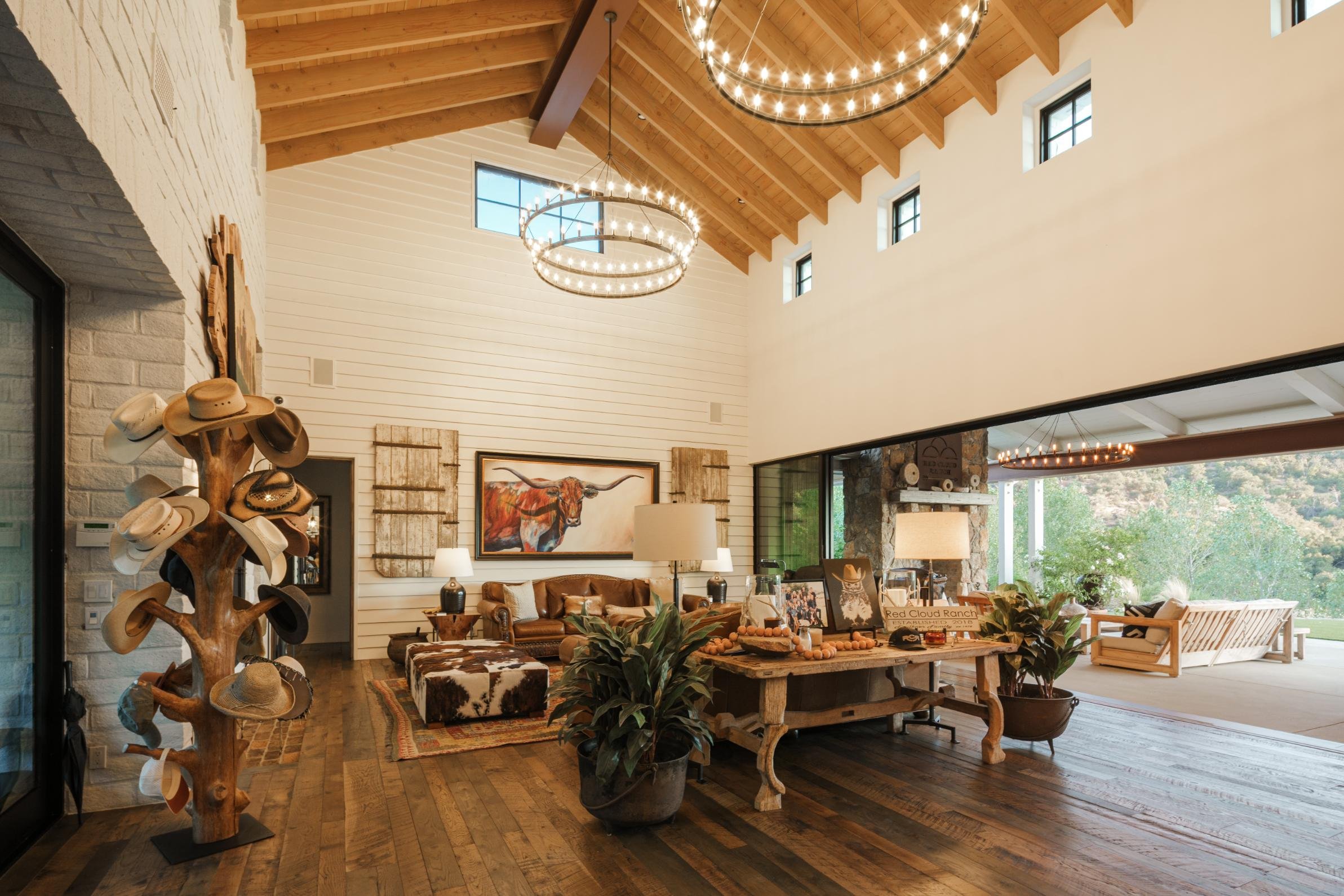Spacious living room with high wooden vaulted ceiling, large windows, and rustic decor. Furniture includes a leather sofa, cowhide ottoman, and a wooden table with plants. Unique tree-shaped hat stand with hats. Artworks on the walls, including a ste