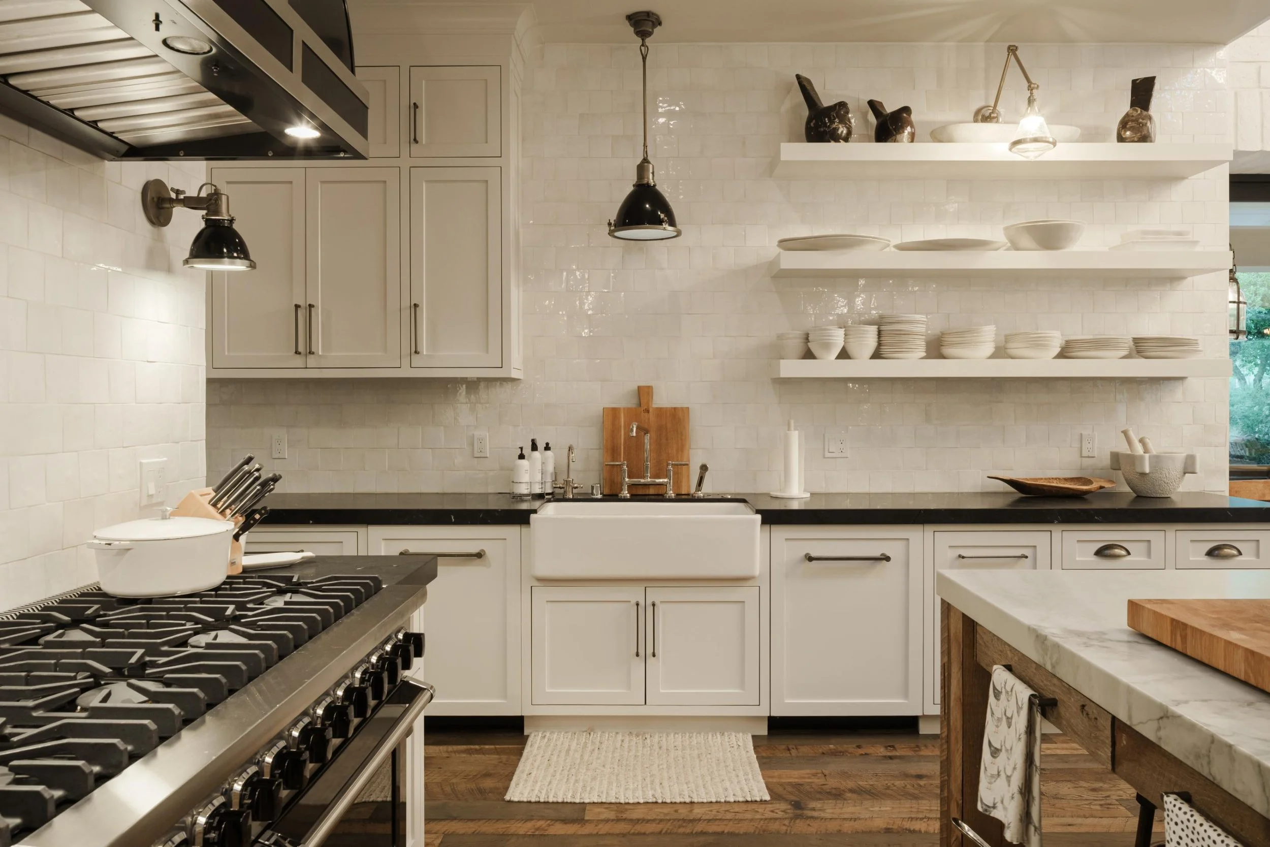 A modern kitchen with white cabinetry, a farmhouse sink, open shelves with dishes and decorative vases, a gas stove, and a kitchen island with a marble countertop.