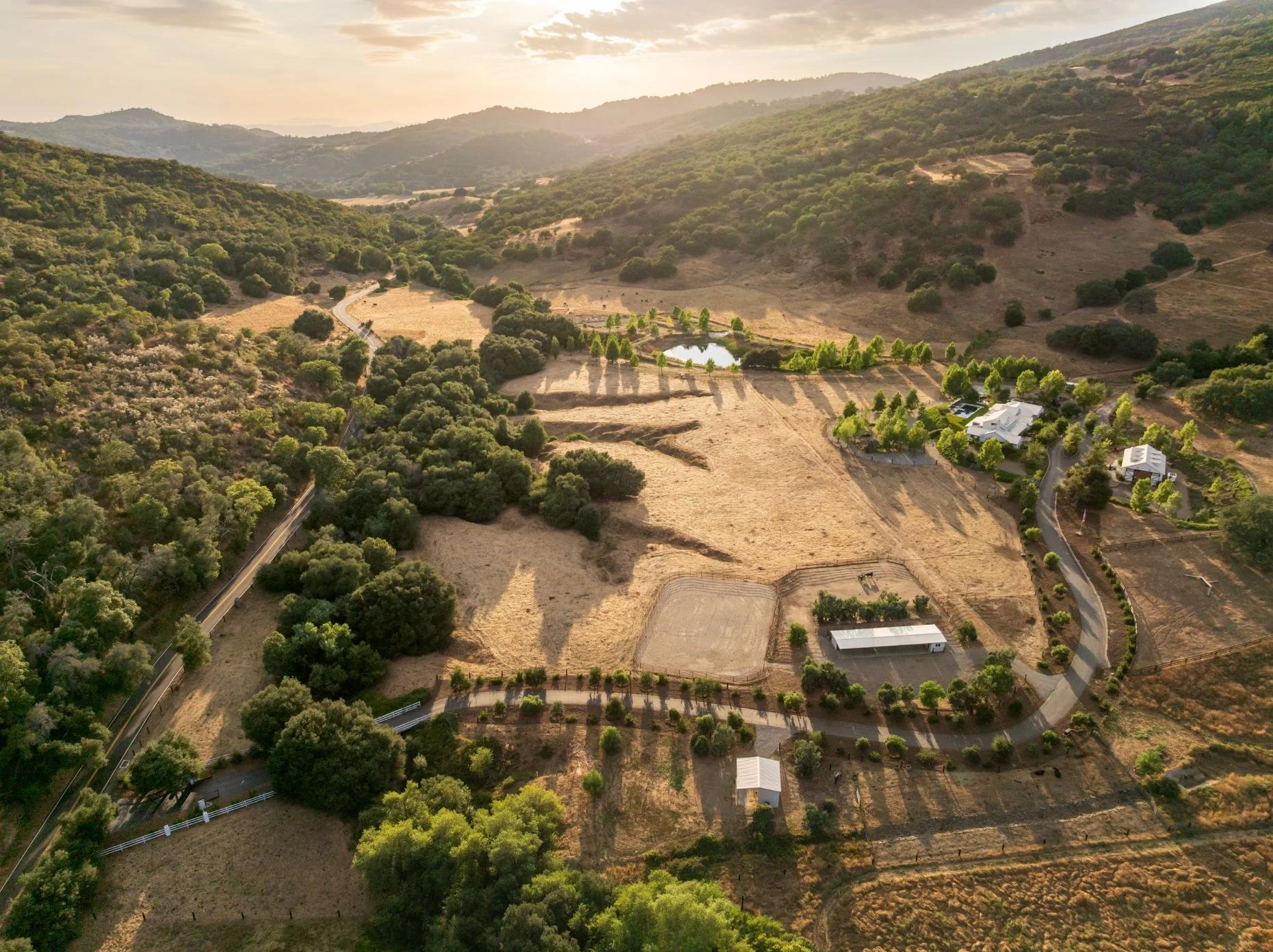 Aerial view of a rural landscape with rolling hills, scattered trees, a pond, and a few buildings along winding roads.