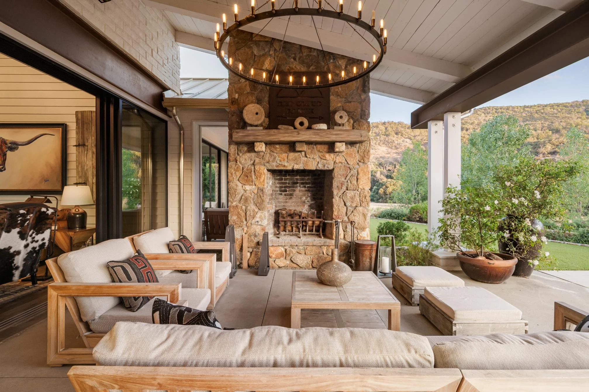 Outdoor living space with stone fireplace, wooden furniture, potted plants, and a view of green landscape and hills.