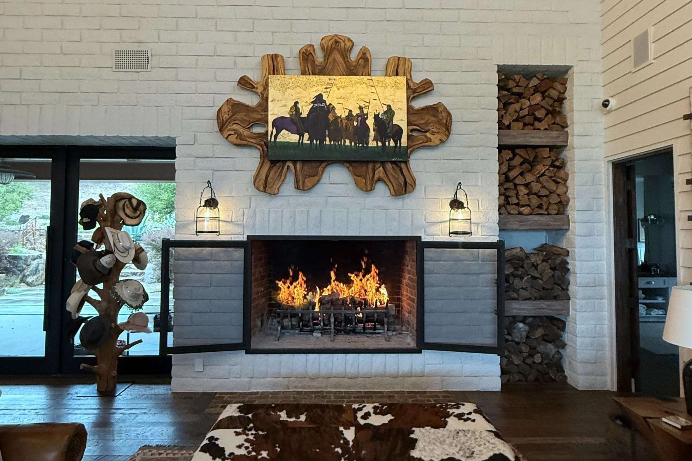 Living room with a white brick fireplace and a wood carved frame above it, featuring a Western painting. To the left, a tree-shaped hat rack with hats and other items, and to the right, stacked firewood in built shelves. Two wall sconces flank the fi