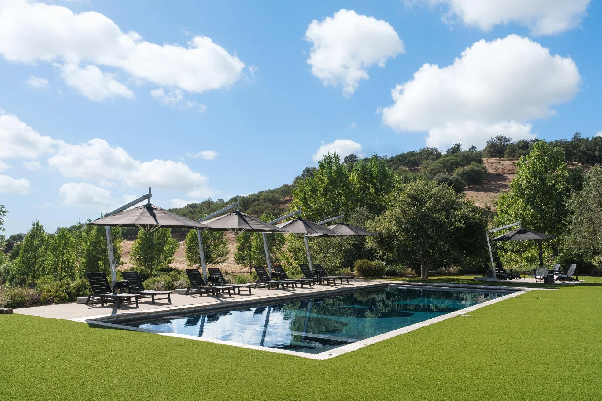 An outdoor swimming pool with black lounge chairs and umbrellas, surrounded by a grassy area, trees, and hills under a partly cloudy sky.