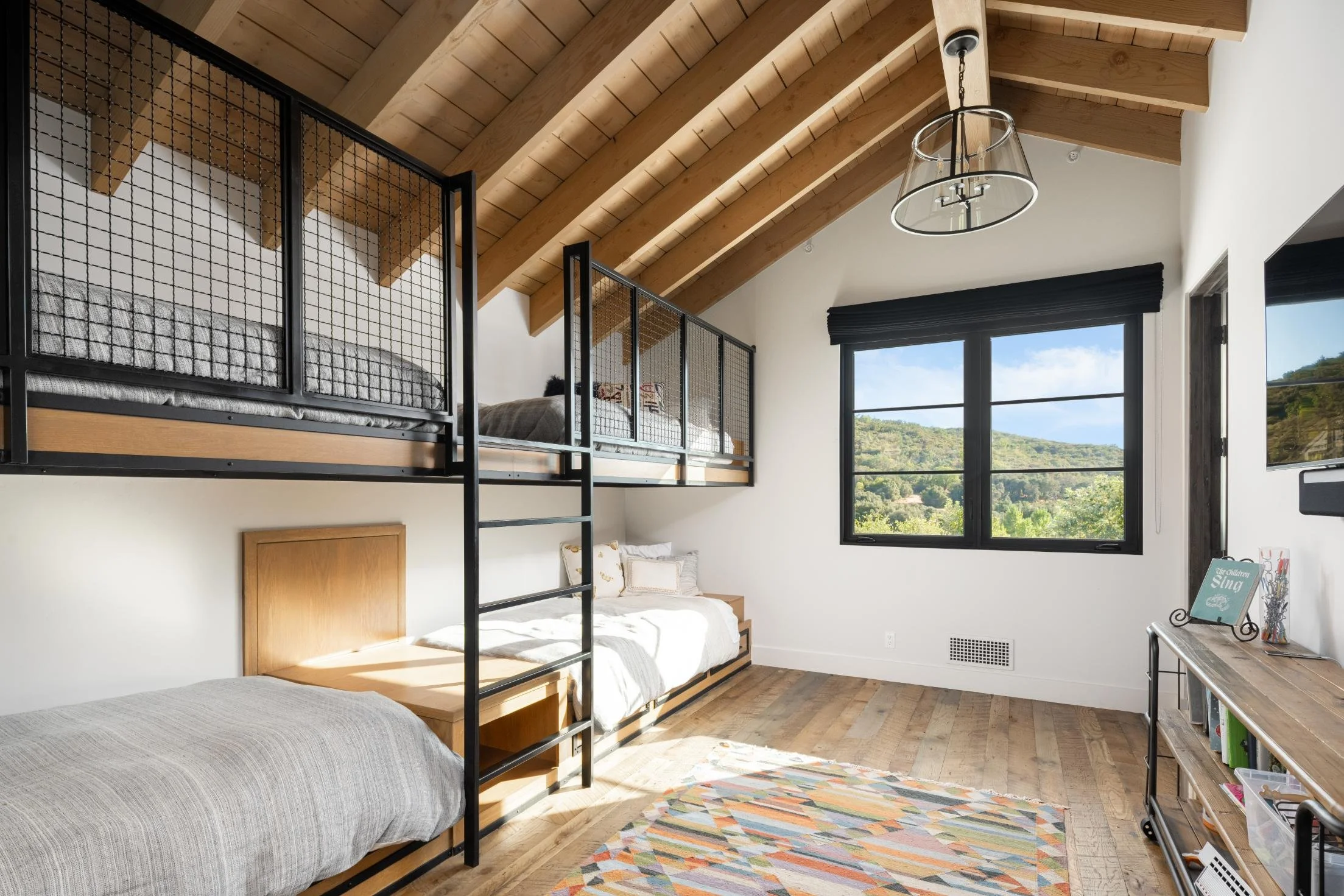 Bedroom with a bunk bed and a lower twin bed, wooden ceiling, large window showing greenery outside, wooden floor, and a bookshelf against the wall.