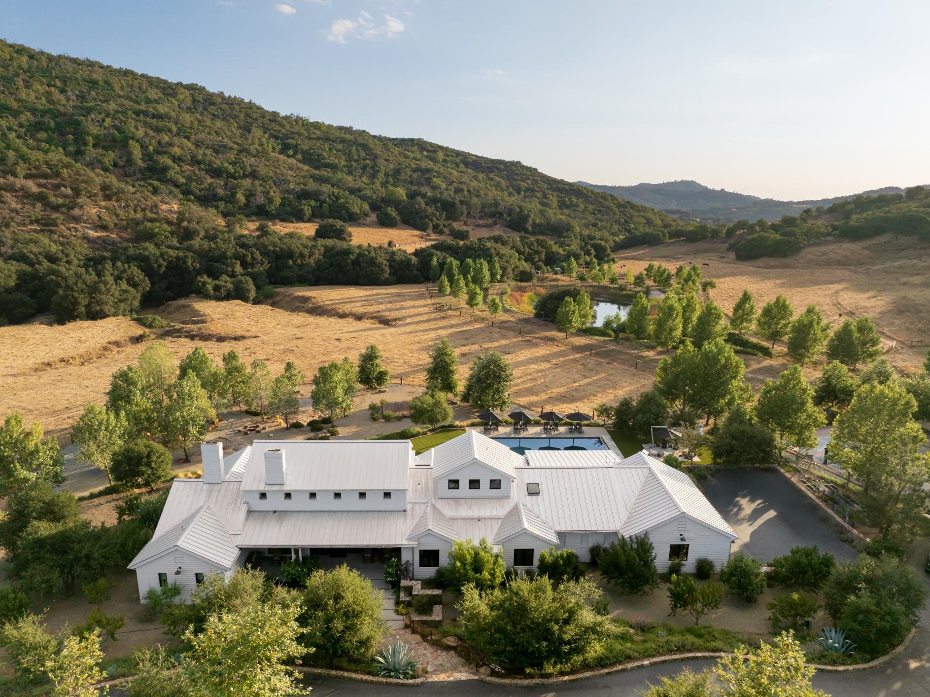 A white house with a metal roof, surrounded by trees, with a swimming pool, set in a rural landscape with hills and fields.