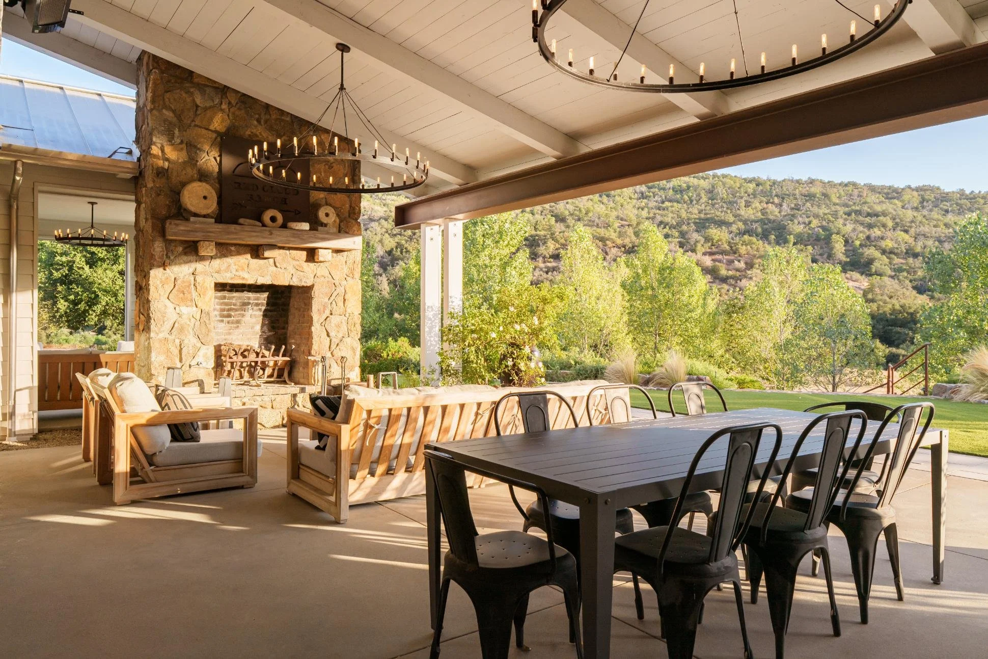 Covered outdoor patio with wooden and metal furniture, stone fireplace, and scenic view of trees and hills in the background, illuminated by chandeliers.