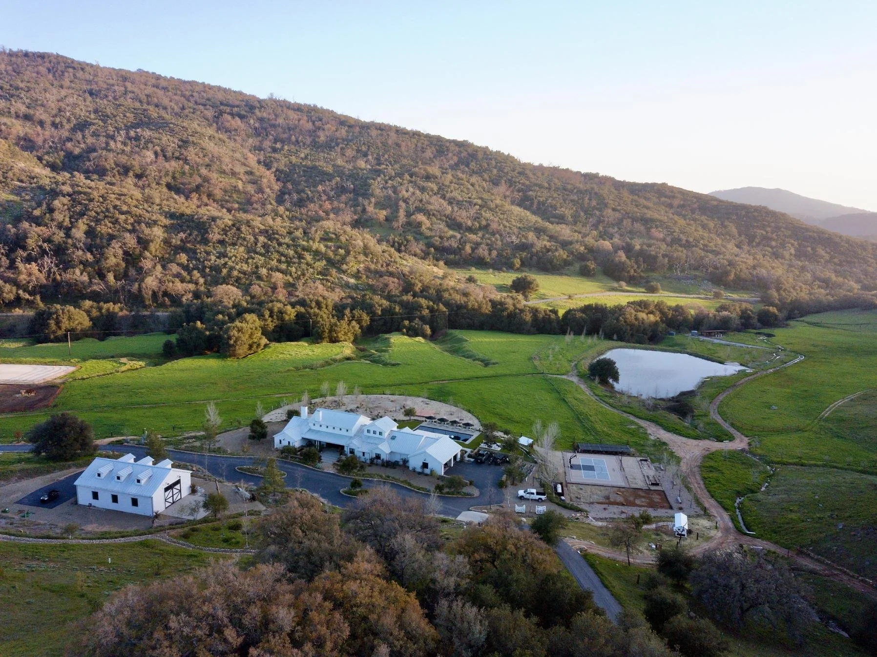 A farmstead with white buildings, including a barn and house, surrounded by green fields and trees, with a pond nearby, set against rolling hills or mountains in the background.