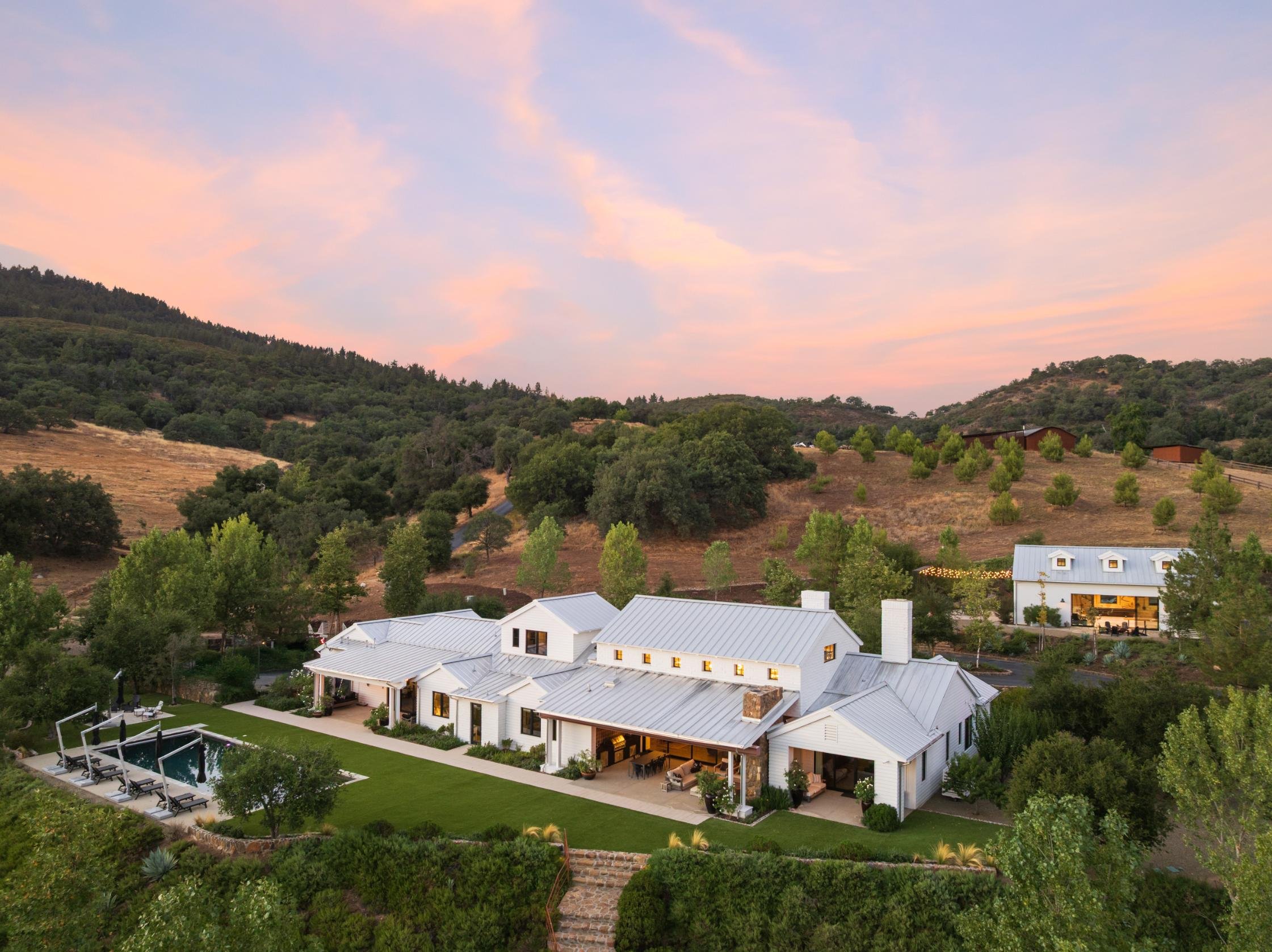 A white house with a metal roof and chimneys, surrounded by trees and a well-maintained lawn with a swimming pool and poolside loungers, set in a hilly landscape at sunset.