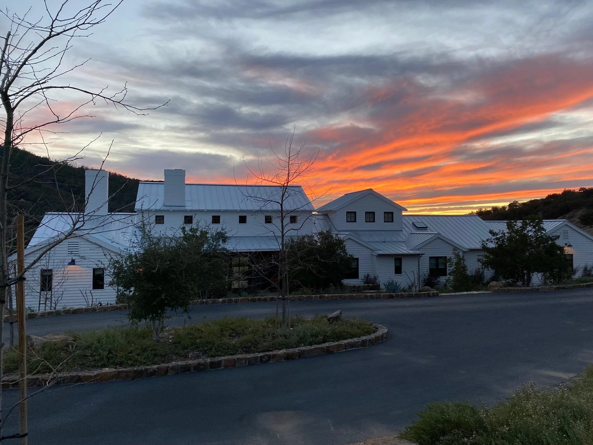 White building with a metal roof at sunset, with a colorful sky of orange and gray clouds, trees, and a curved driveway in the foreground.