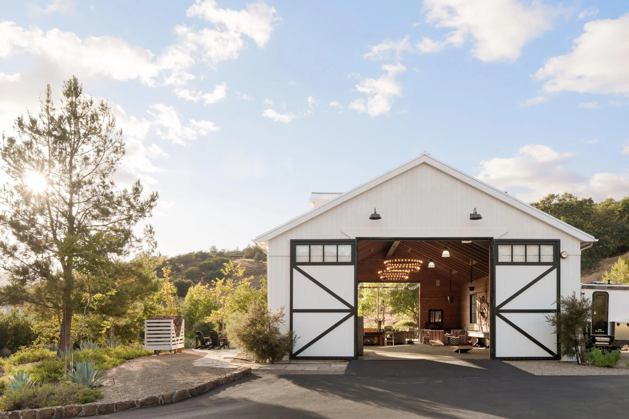 A white barn-style building with open black sliding doors, showcasing a rustic, cozy interior, surrounded by trees and a clear sky with clouds.