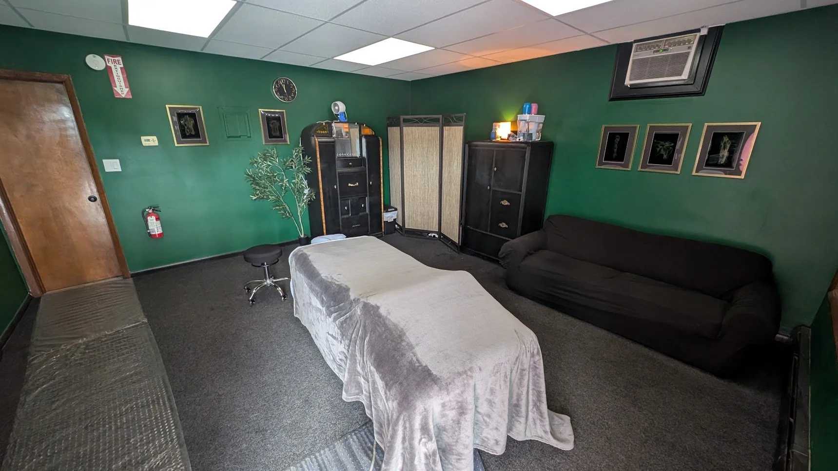 Room with green walls, featuring a massage table with a beige cover, a black sofa, a black chair, a folding screen, framed artwork, a clock, a small plant, and various small items on shelves.