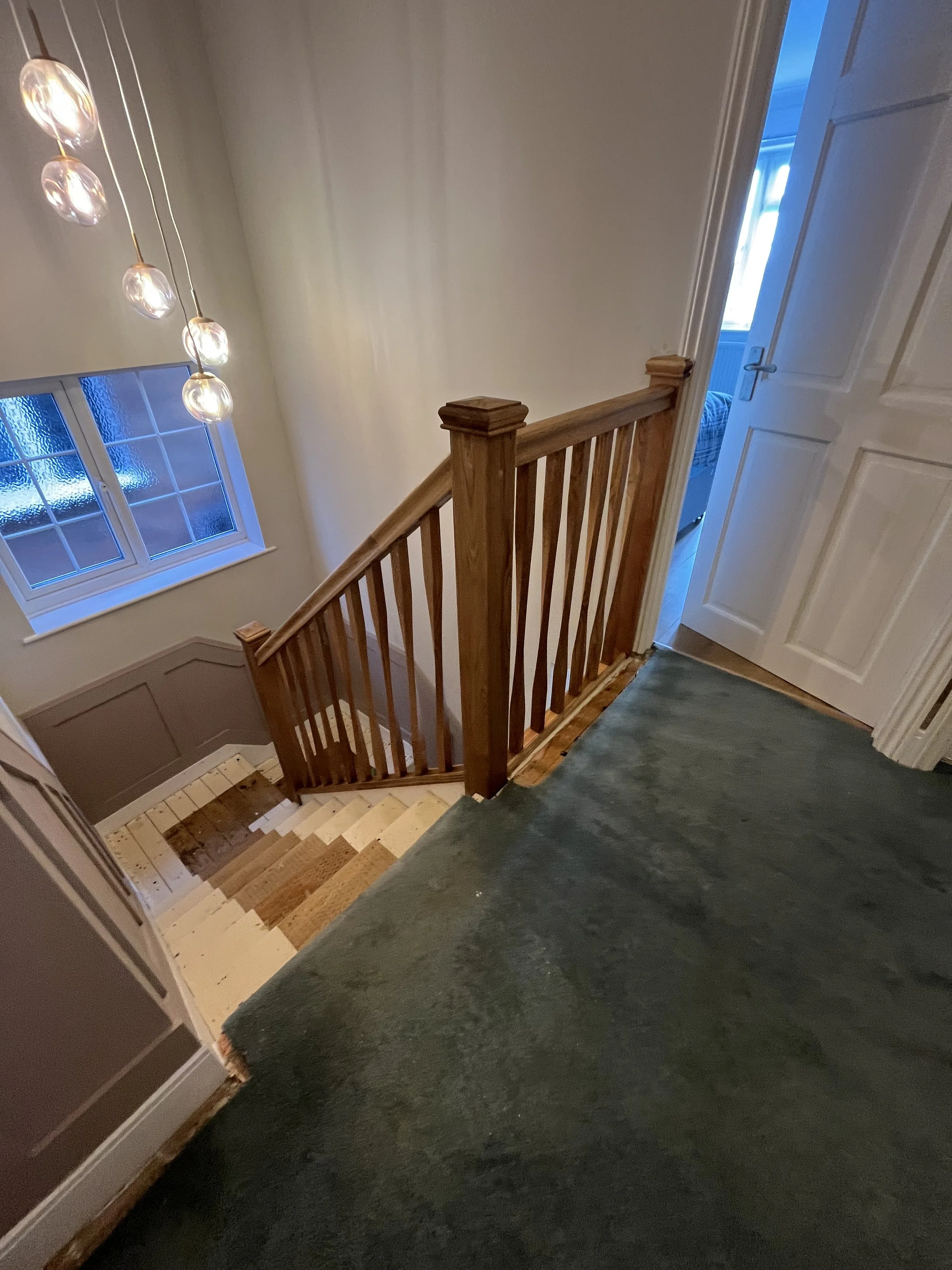 Interior view of a wooden staircase with new railing and a small door underneath. Large windows show an outdoor backyard with green grass and a wooden fence.