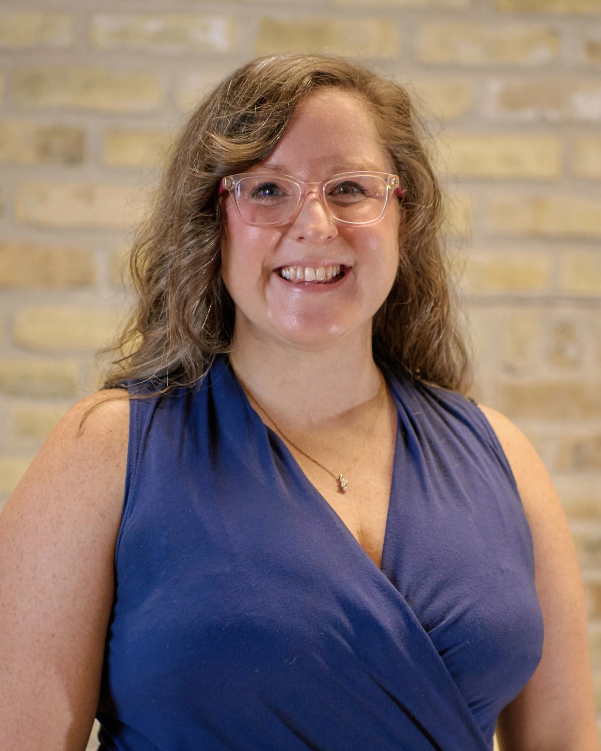 A woman with shoulder-length gray hair, wearing clear glasses with pink accents, a sleeveless maroon ribbed turtleneck top, small earrings, and a gold necklace, smiling in an indoor setting with blurred background.