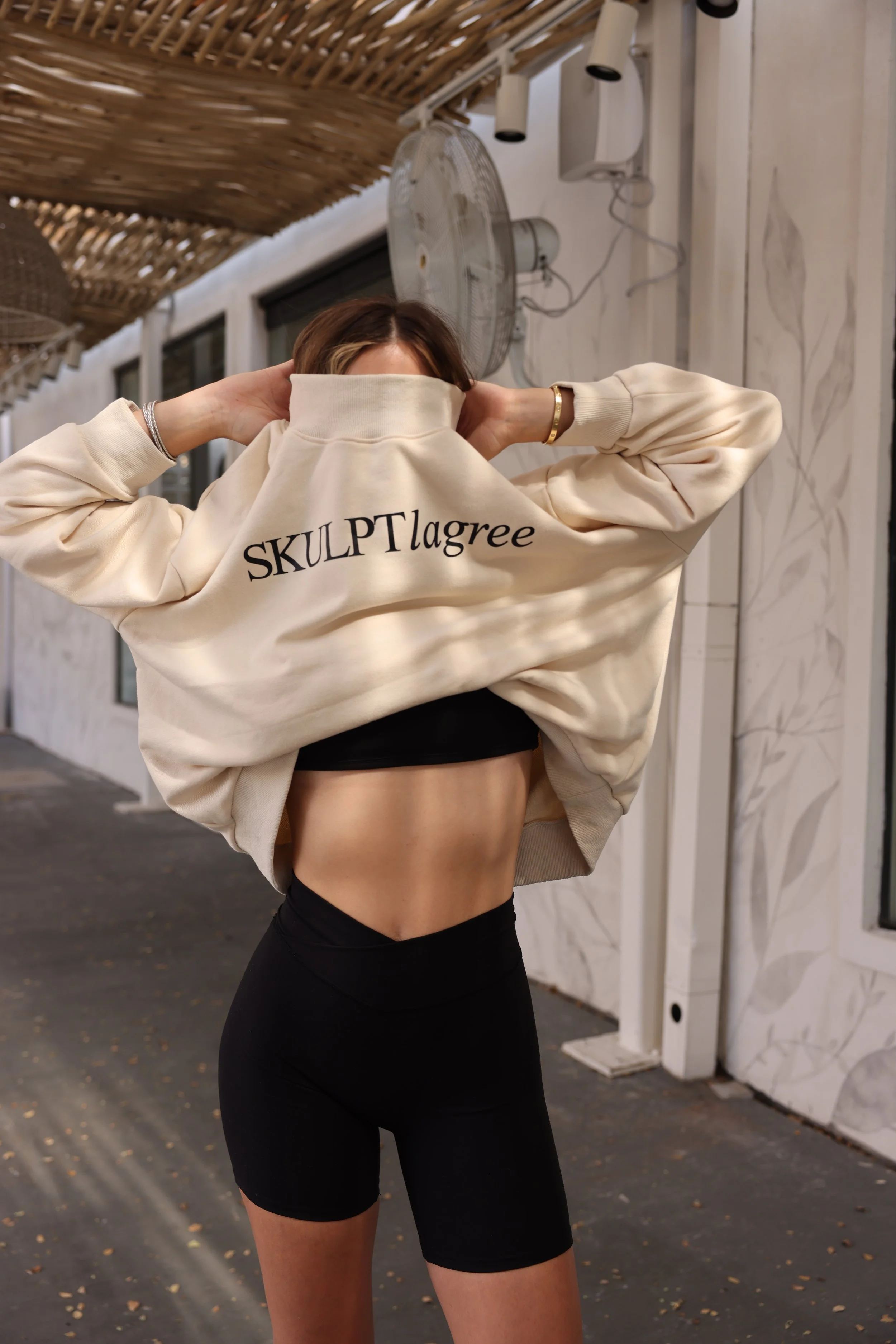 A woman lifting her sweatshirt to reveal her toned abdomen, standing outdoors at night, wearing black sports bra and high-waisted black shorts, with a fan and wall decorations in the background.