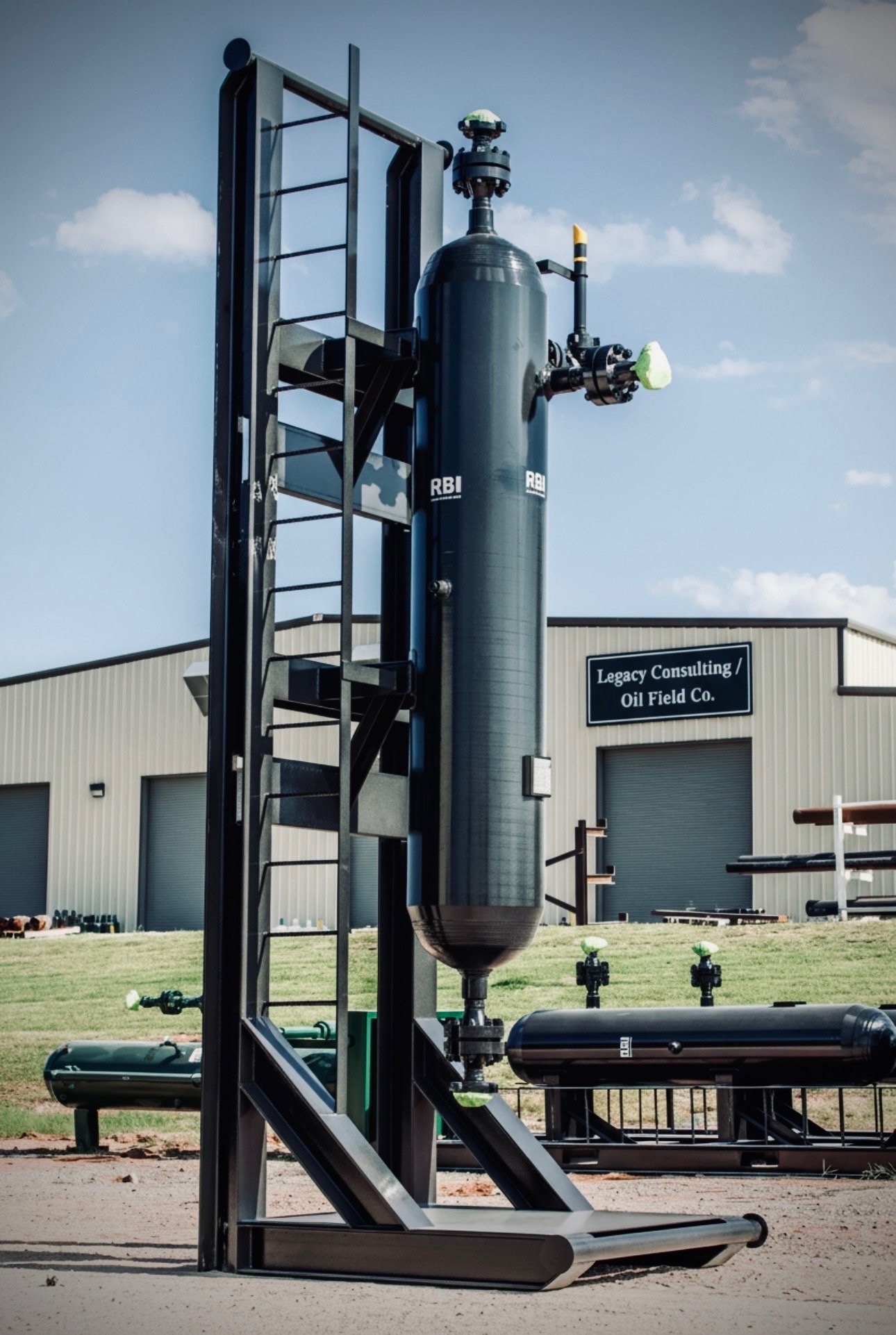 Large vertical industrial equipment with pipes and gauges, situated outside a building with a sign that reads 'Legacy Consulting / Oil Field Co.' under a partly cloudy sky.