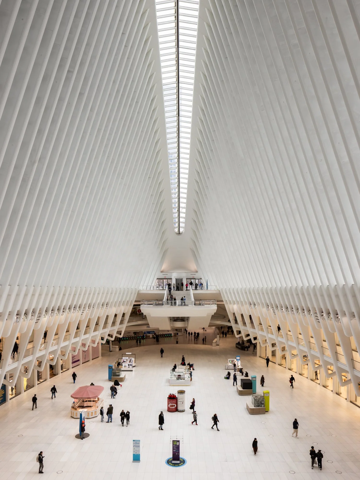Interior of the Oculus at World Trade Center Transportation Hub in New York City, showcasing its modern, white, ribbed architectural design with high ceilings and a skylight, populated with visitors and retail kiosks.