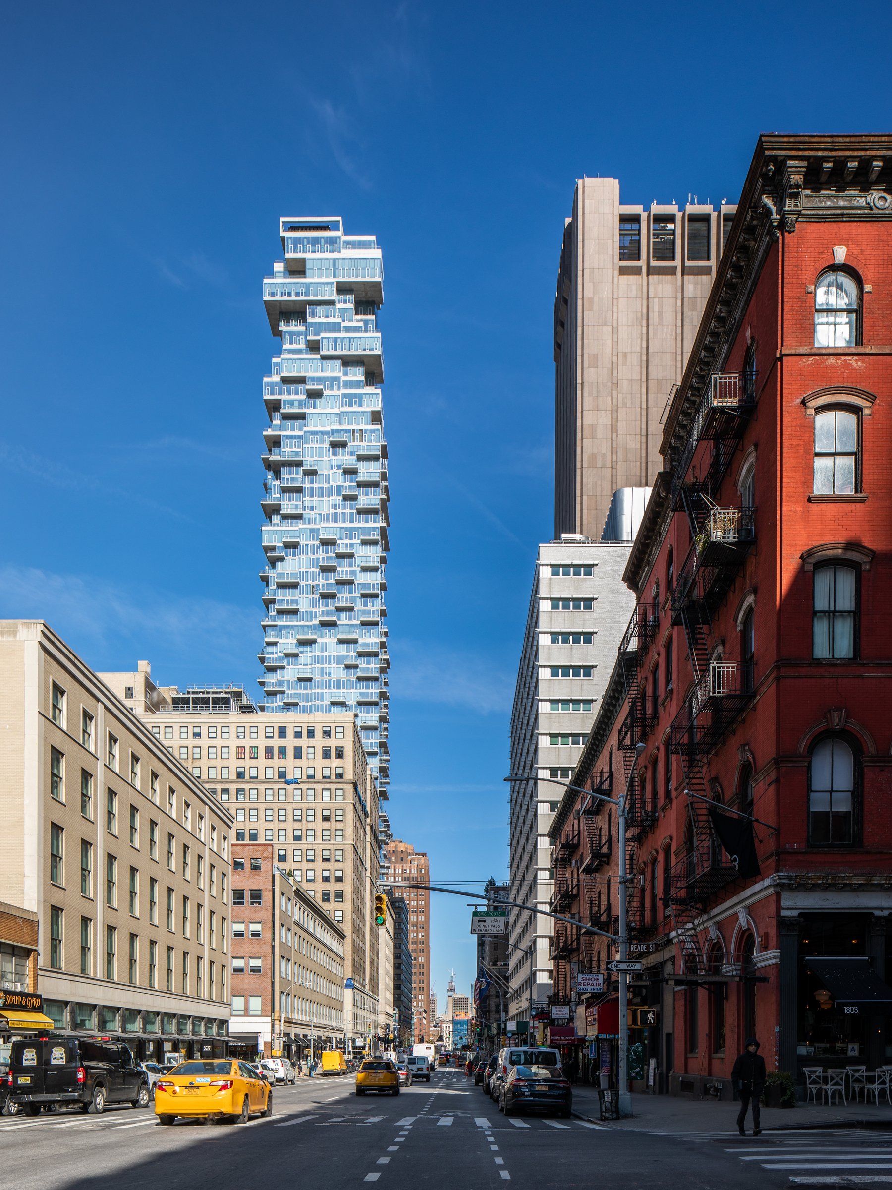 City street with parked cars and yellow taxis, tall modern and historic buildings, blue sky with a few clouds.