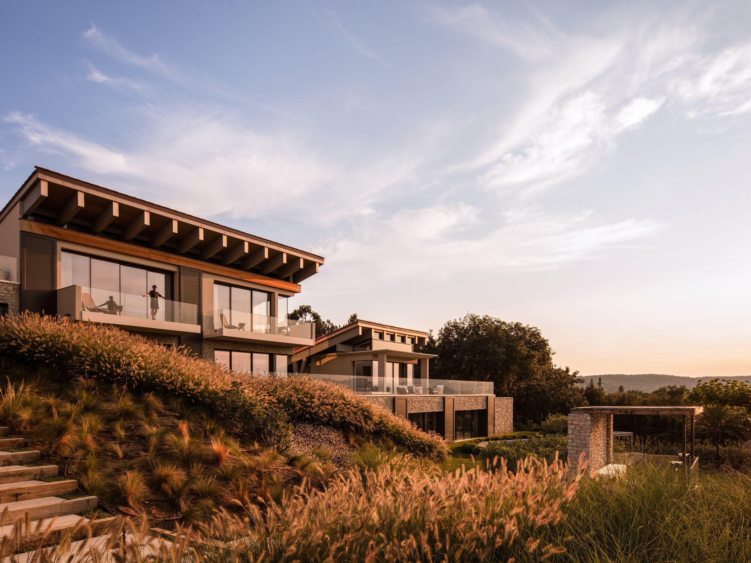 Modern multi-level house on a hillside with large glass windows, balconies, and landscaped garden with grasses, trees, and a stone structure, during sunset.