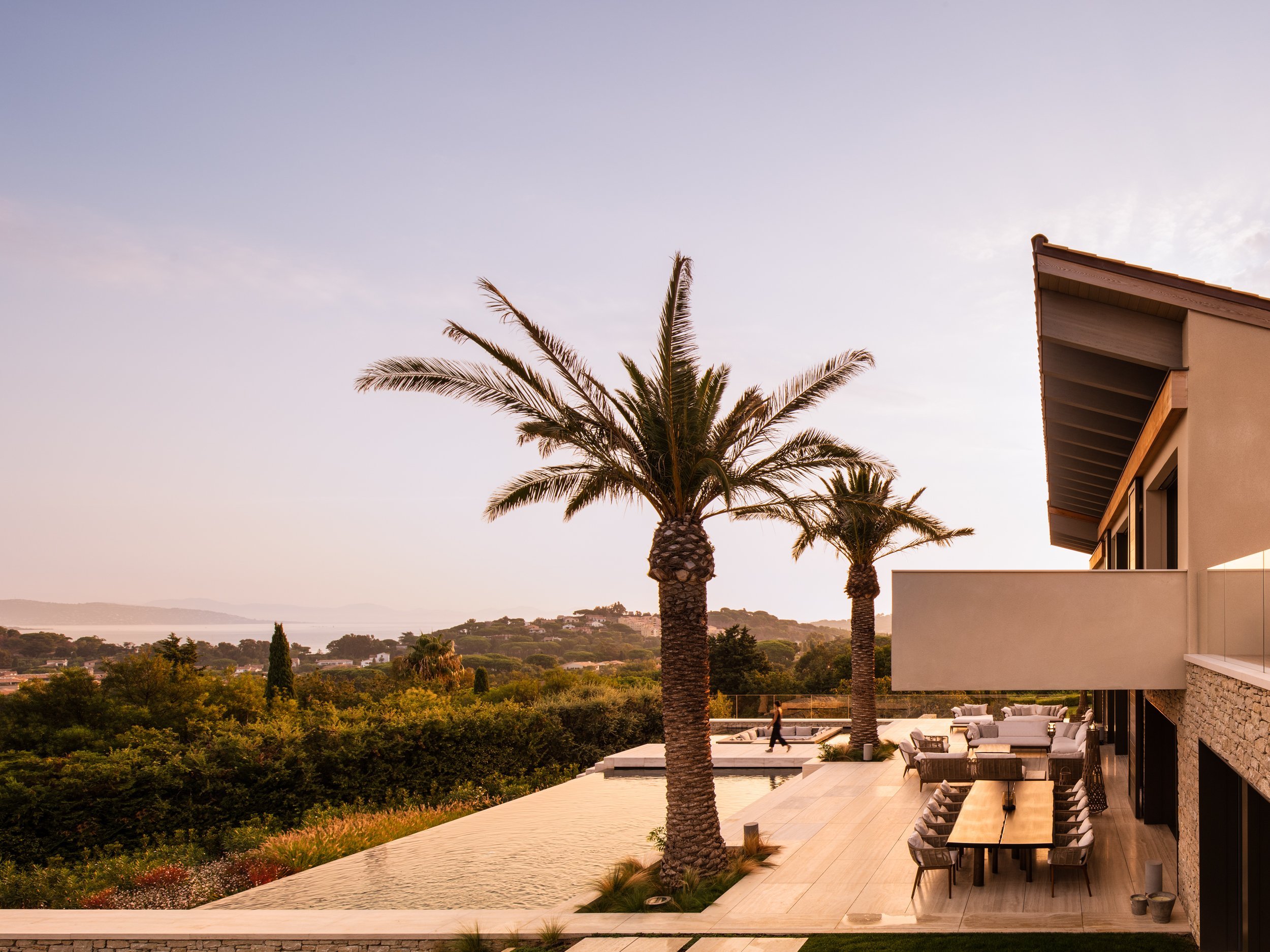 Luxury modern house with outdoor dining area, tall palm trees, and lush green landscape at sunset.