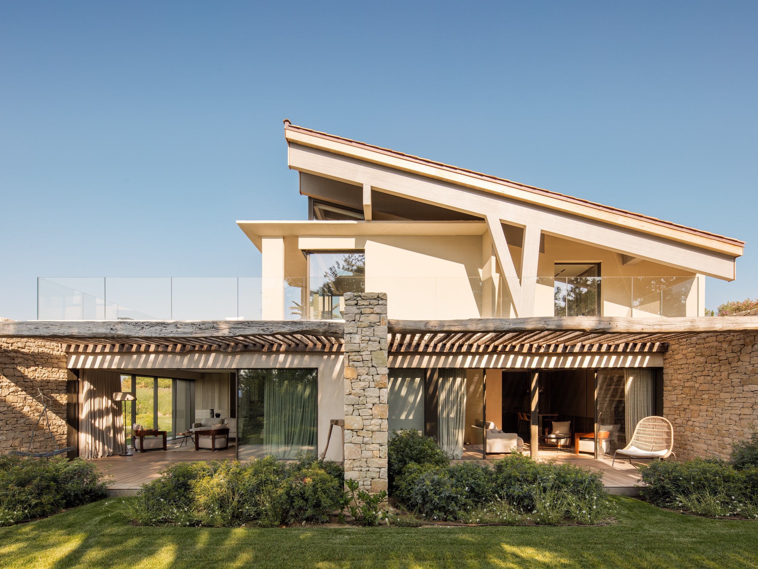 Modern multi-level house with stone and stucco exterior, large glass sliding doors, and outdoor seating area, under a clear blue sky.