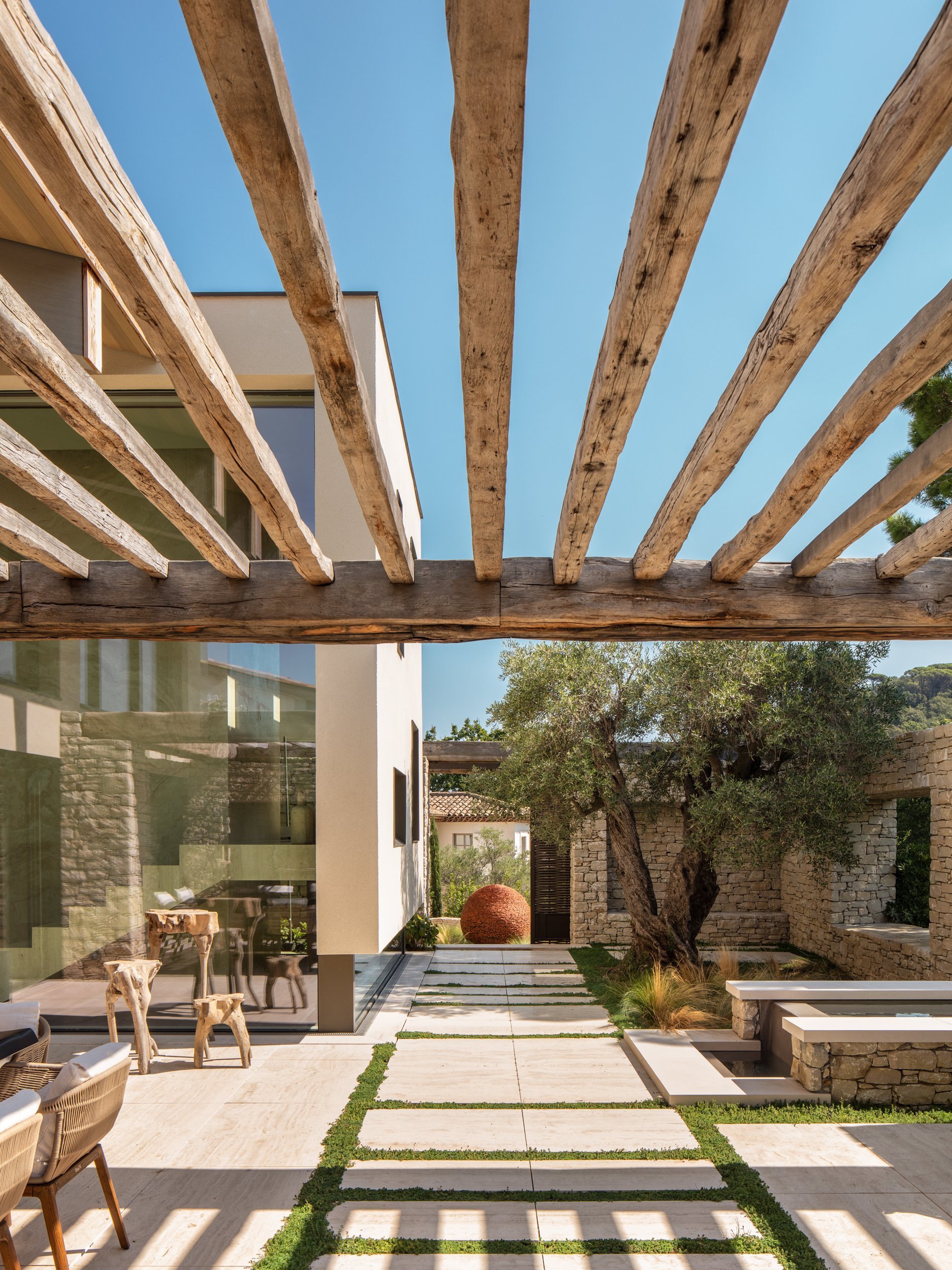 Modern outdoor patio with stone pathway, trees, and a wooden pergola with a clear blue sky.