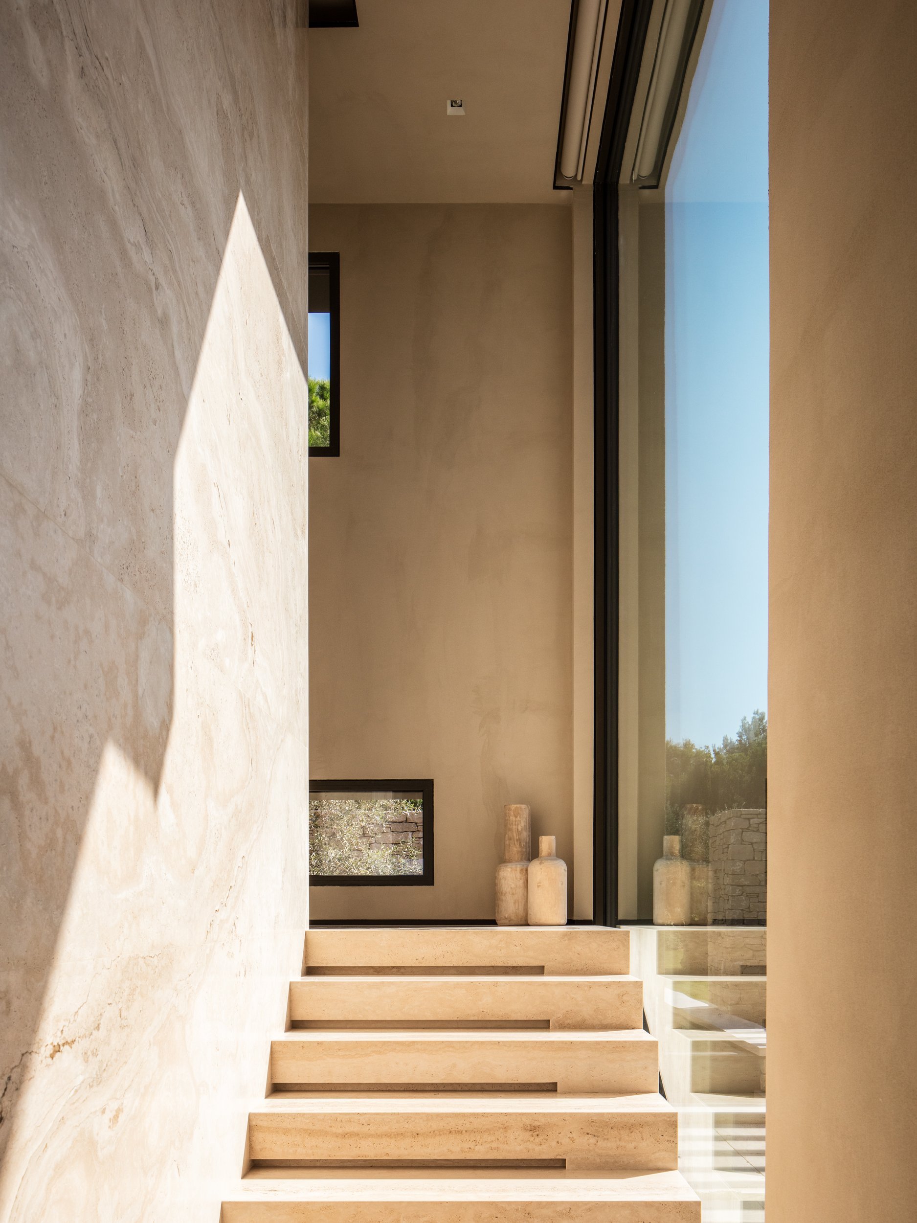 Minimalist interior featuring a staircase with wide steps, natural-toned walls, and decorative vases near large glass doors leading outside, with sunlight casting shadows.
