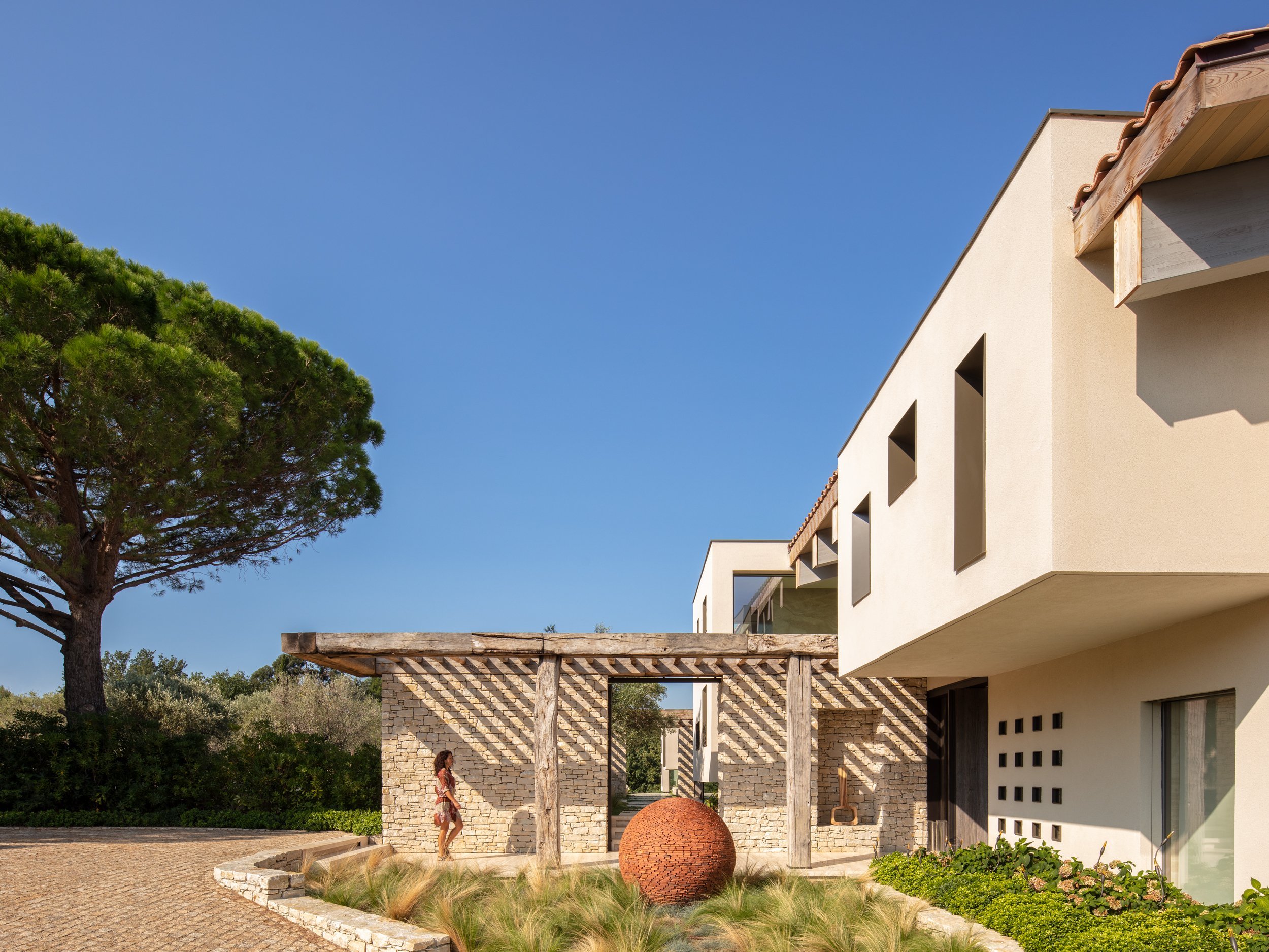 Modern house with white exterior, angular windows, and stone and wood accents, surrounded by greenery under a clear blue sky.