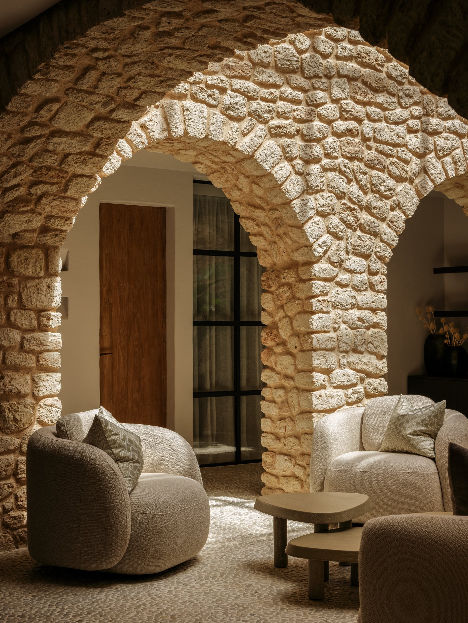 Living room with exposed stone brick arches, beige armchairs with pillows, a small wooden coffee table, a dark sideboard with vases, and a glass door leading outside.